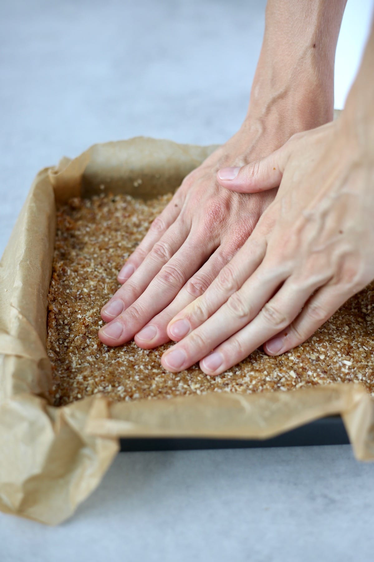 two hands pressing down on some granola bar dough that is inside of a baking dish lined with brown parchment paper