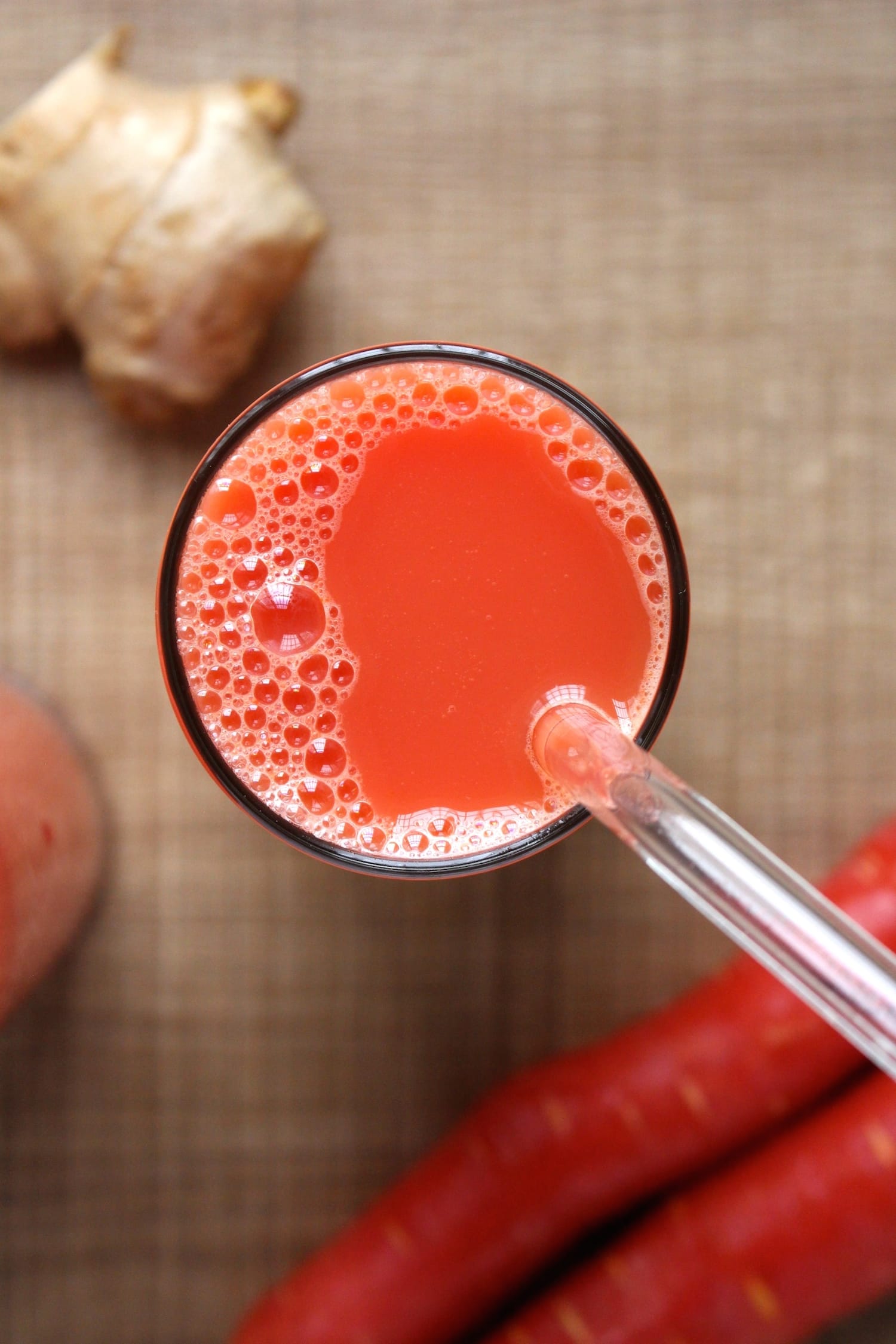 An overhead shot of bubbly apple, carrot and ginger juice made in a blender in a glass with a glass straw poking out. 