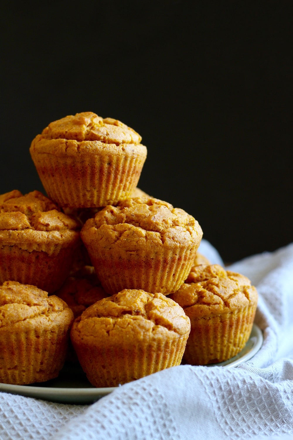 Sunlight shinning on a big stack of vegan pumpkin cornbread muffins stack on a plate with a grey textured towel beneath it. 