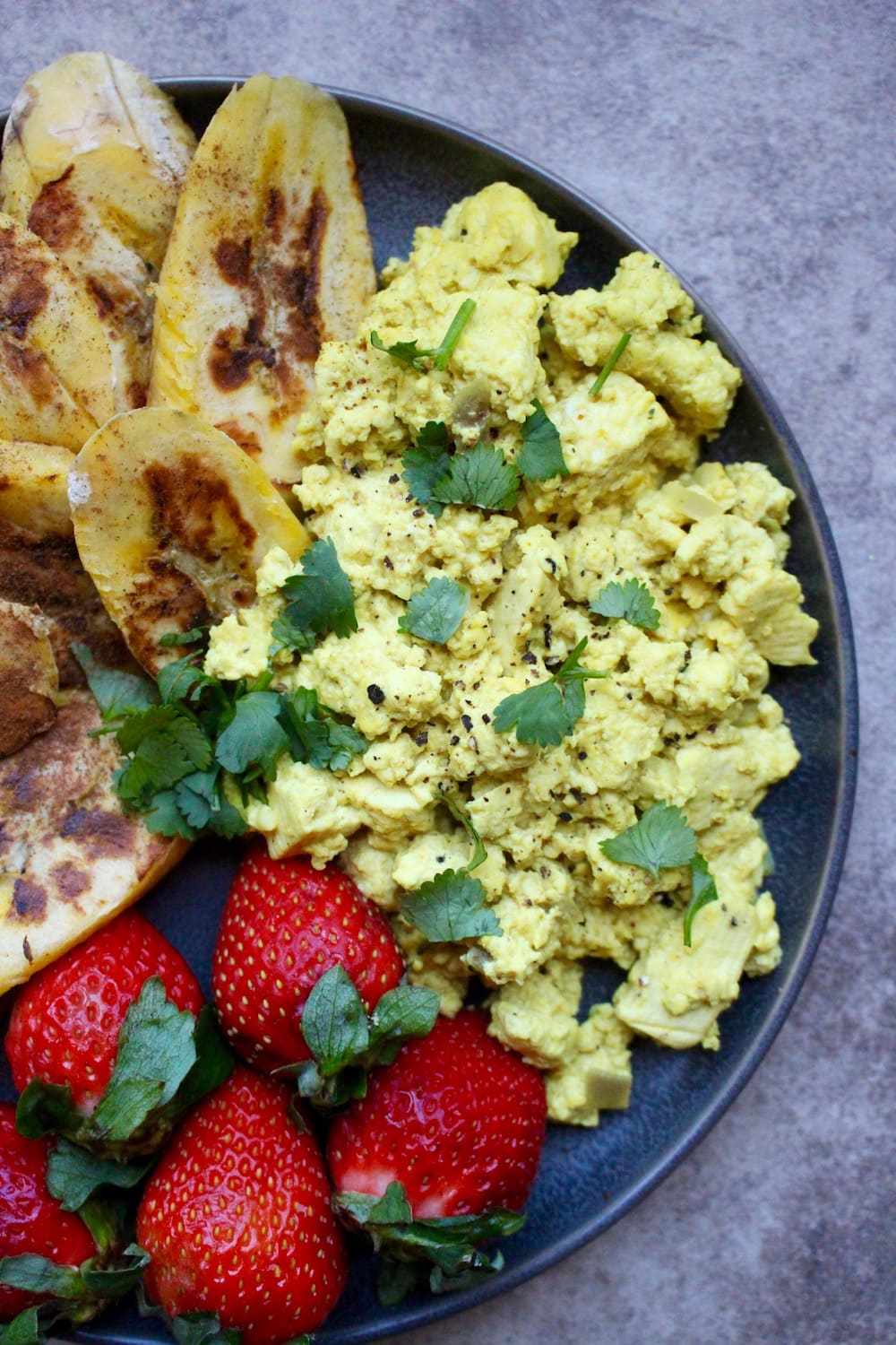 top view of cheesy vegan tofu scramble on a plate with cilantro and strawberries and plantains on the side close up