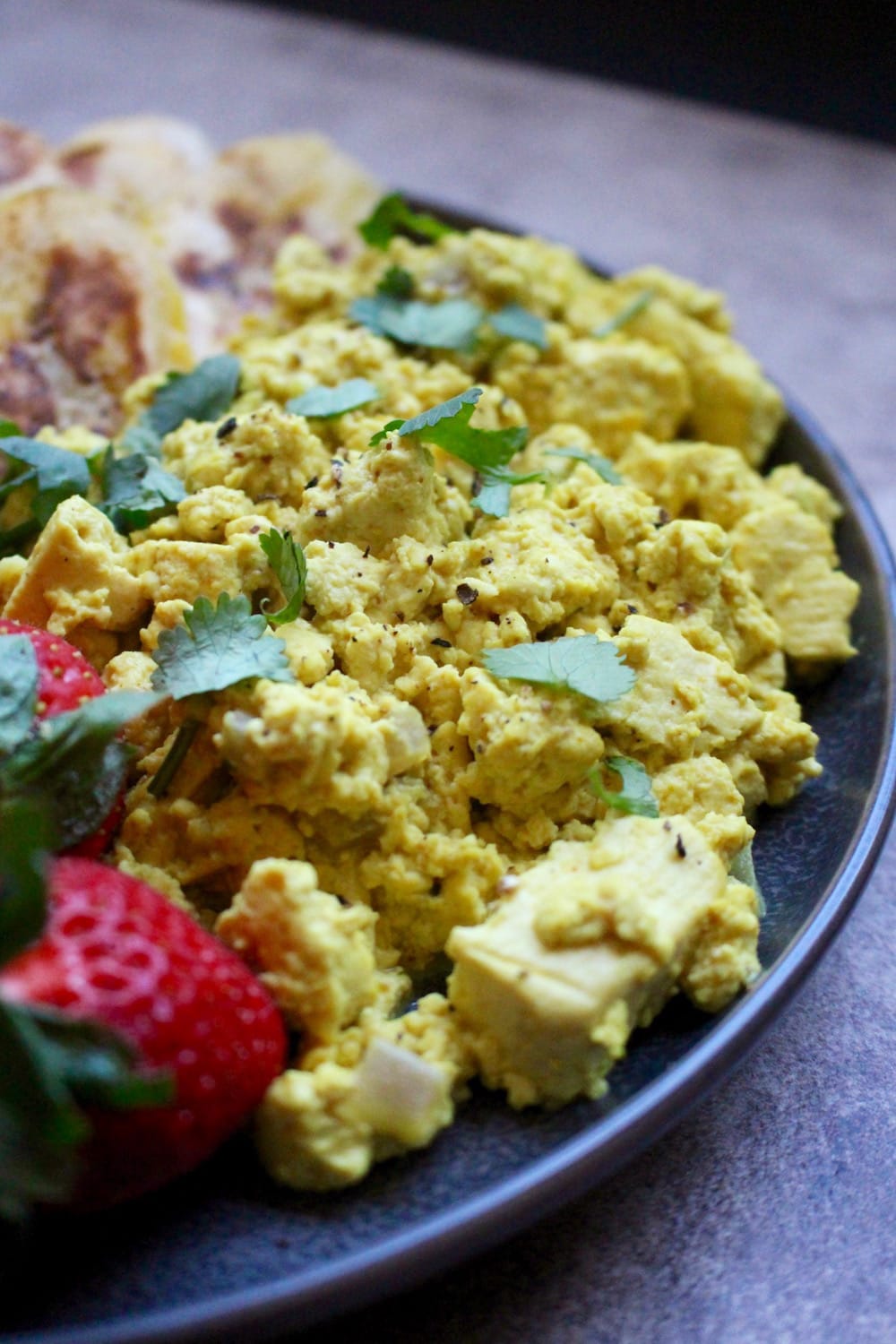 cheesy vegan tofu scramble on a plate with cilantro and strawberries on the side