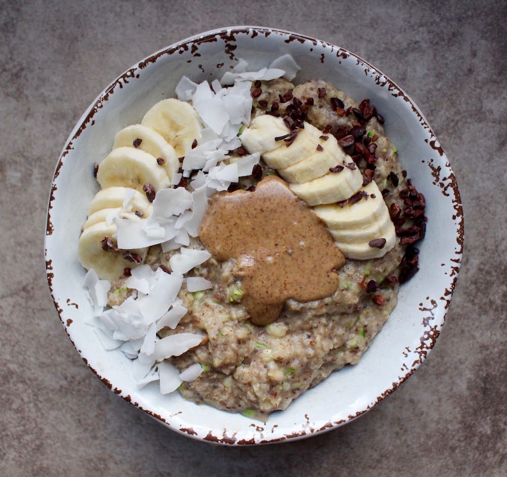 Zoats (a.k.a. oatmeal cooked with shredded zucchini) in a bowl topped with banana slices, coconut flakes, cacao nibs and almond butter