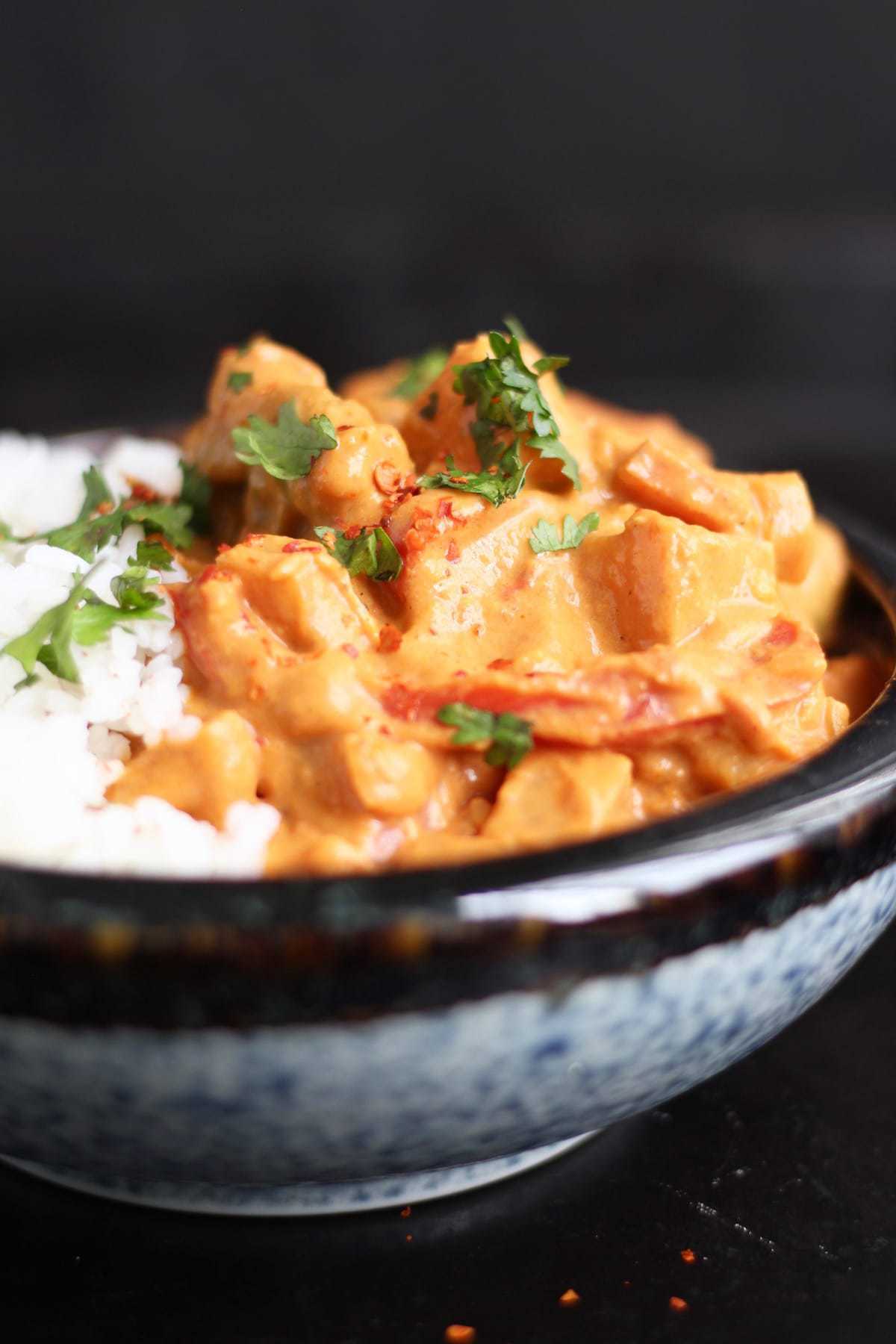 A close up of vibrant orange peanut butter sweet potato curry in a black and blue bowl paired with white rice and green cilantro on top.