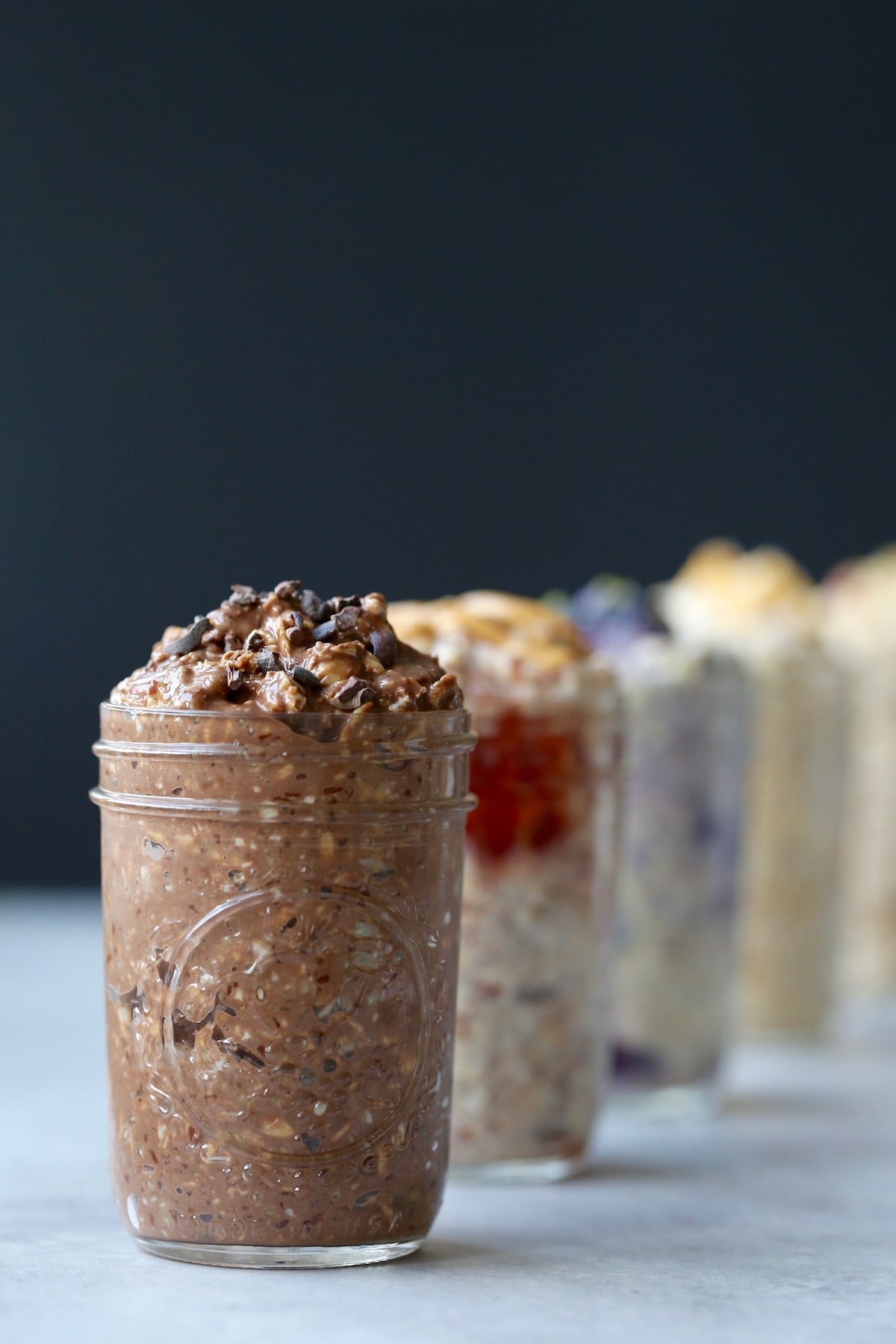 A line of different flavored overnight oats in mason jars with double chocolate in front topped with cacao nibs. 