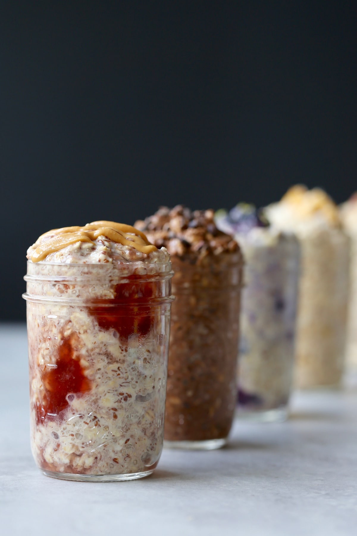 5 mason jars filled with different flavors of overnight oats in a line, with peanut butter and jelly closest to the viewer. 