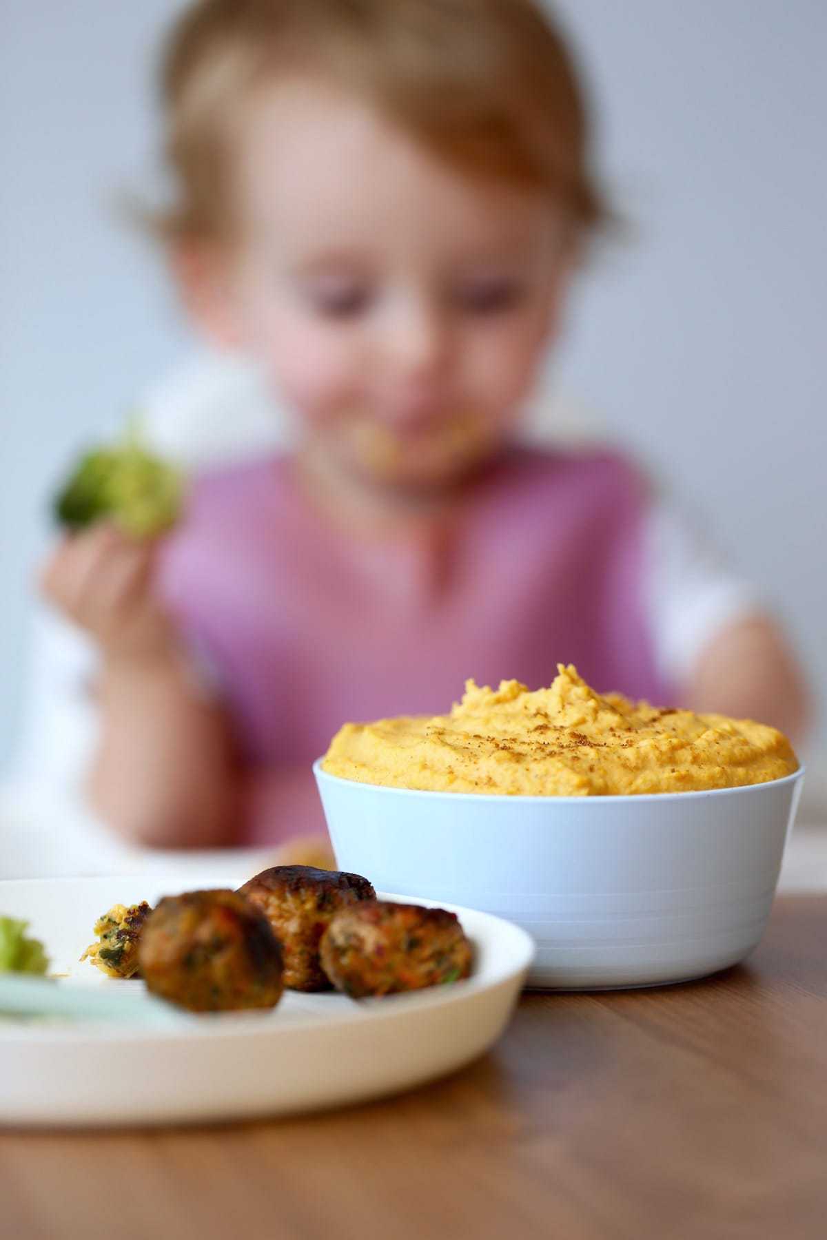 a bowl of carrot hummus on a table with a little girl in the background