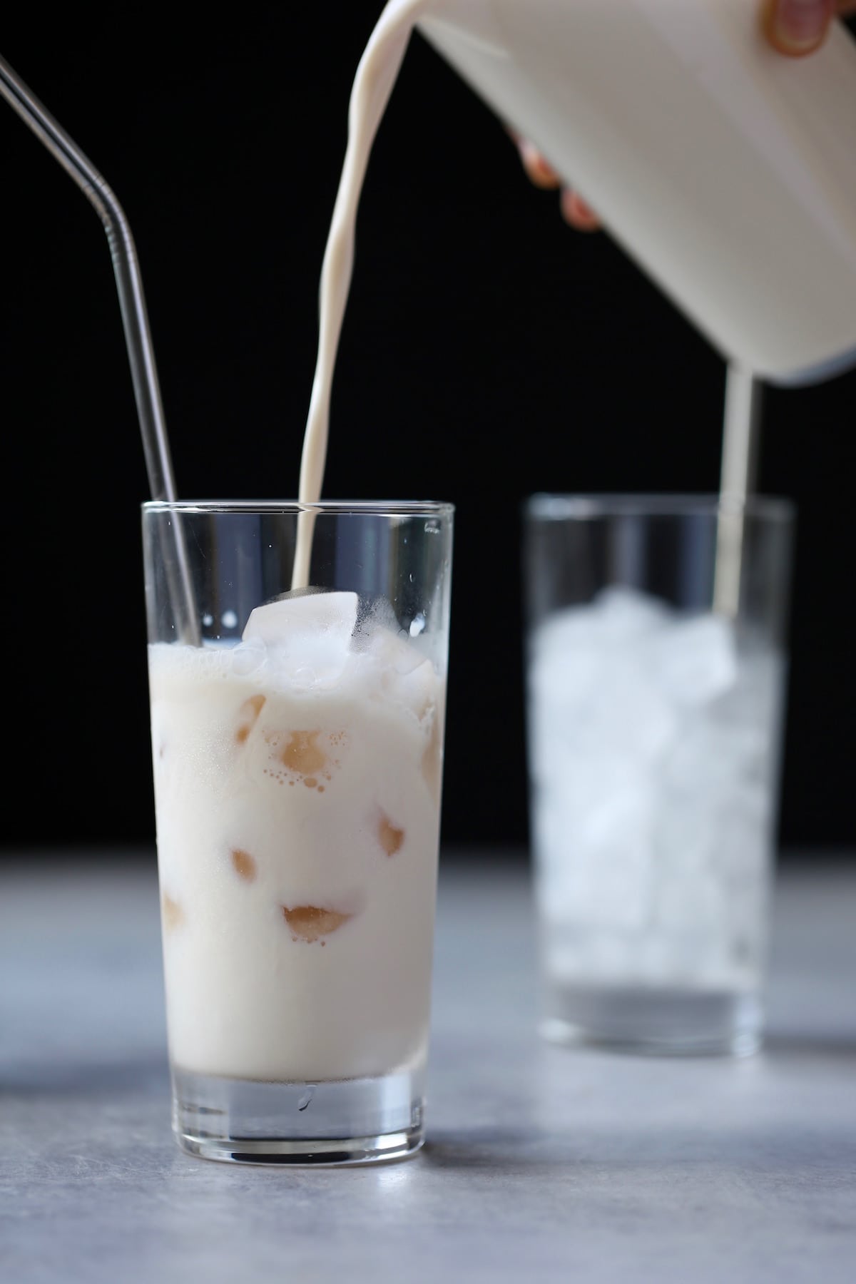 Milk being poured into a tall glass filled with iced and a metal spoon. 