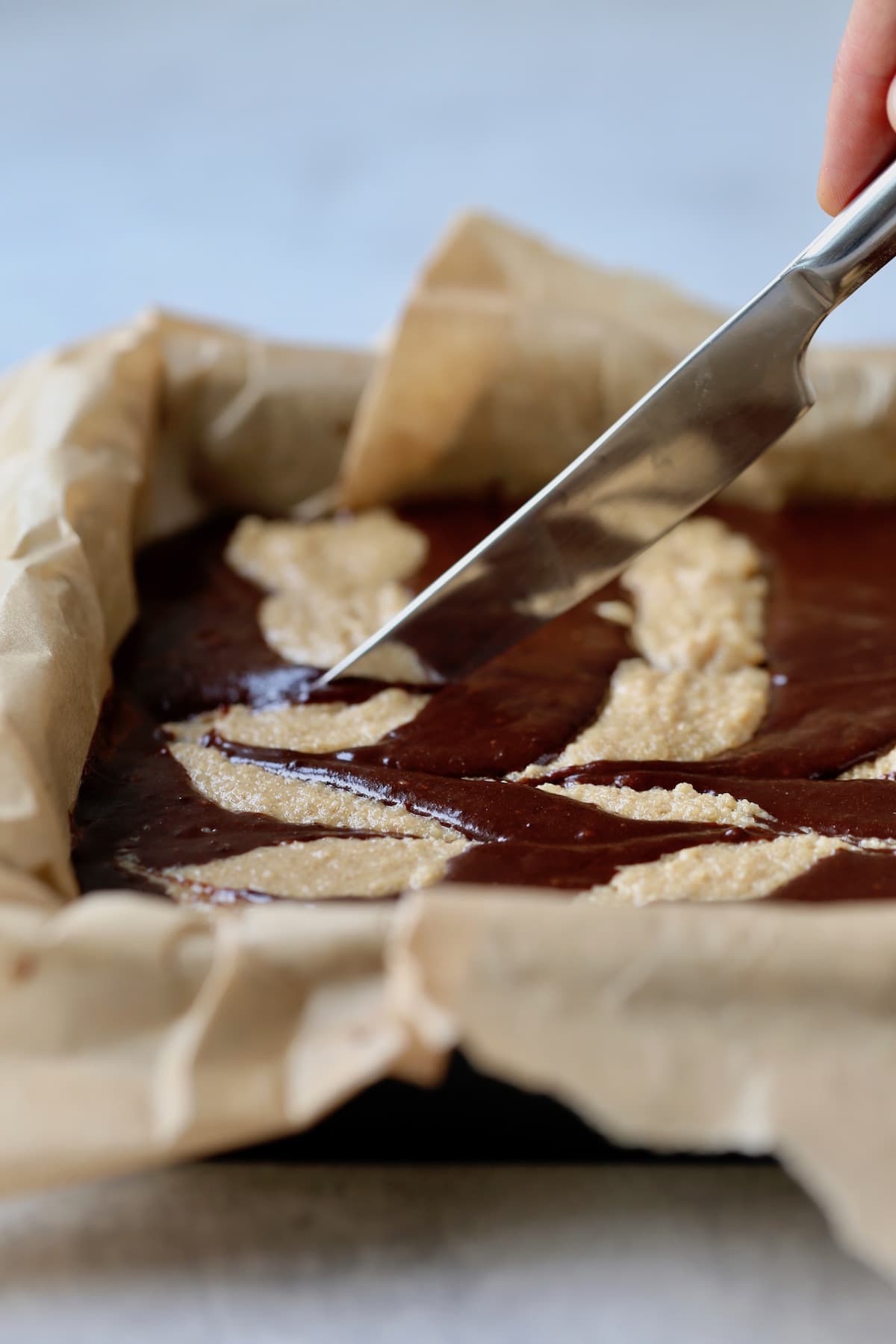 A butter knife being dragged through brownie batter and peanut butter batter that's in a parchment paper lined baking dish. 