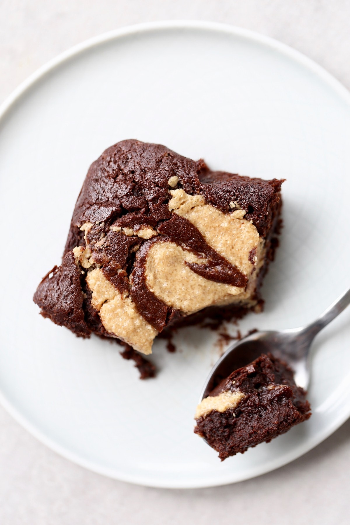 A fudgy vegan brownie with a beautiful peanut butter swirl on a white plate with a spoon scooping up a bite. 