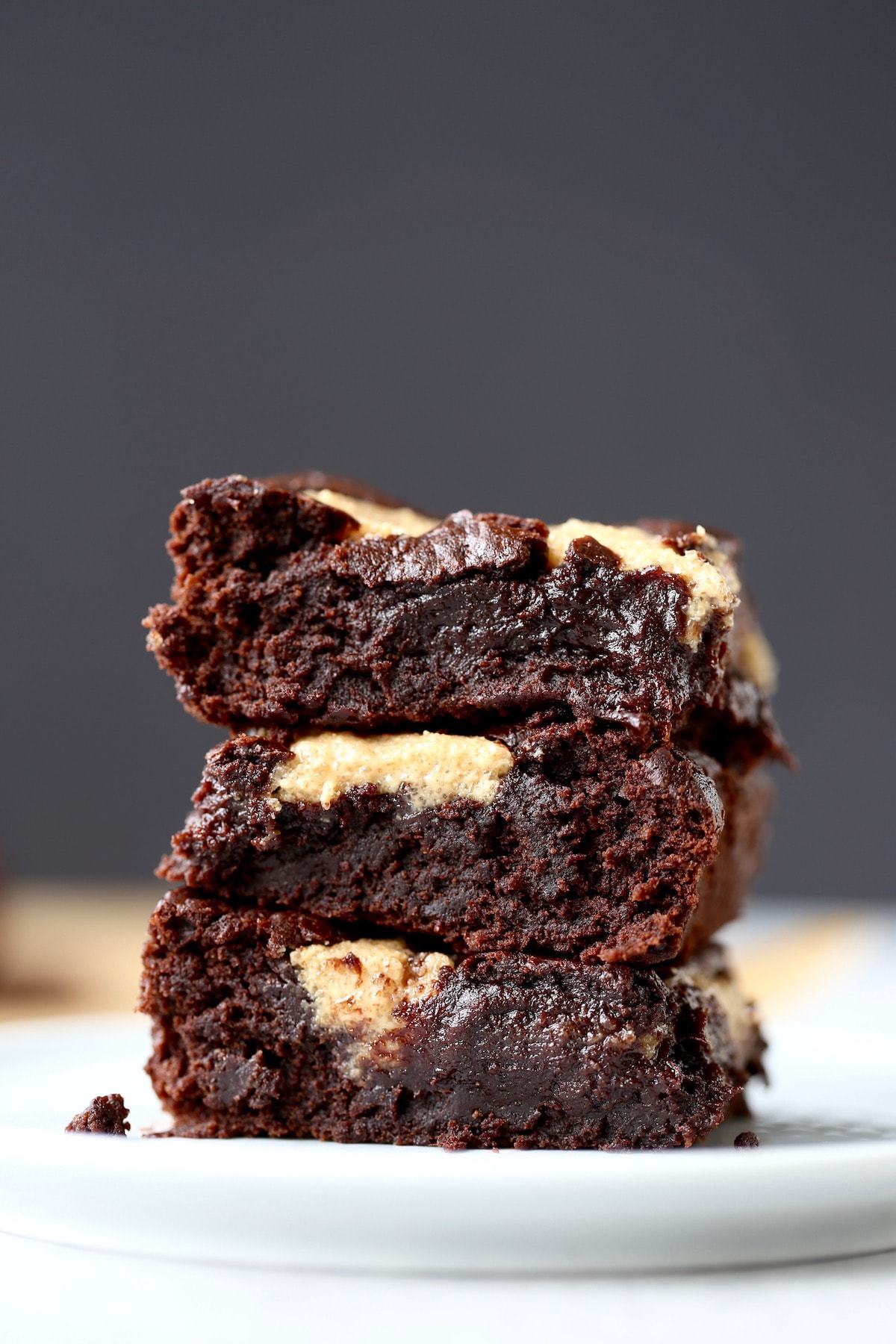 Three fudgy peanut butter topped brownies stacked on each other on a small white plate with a black background. 