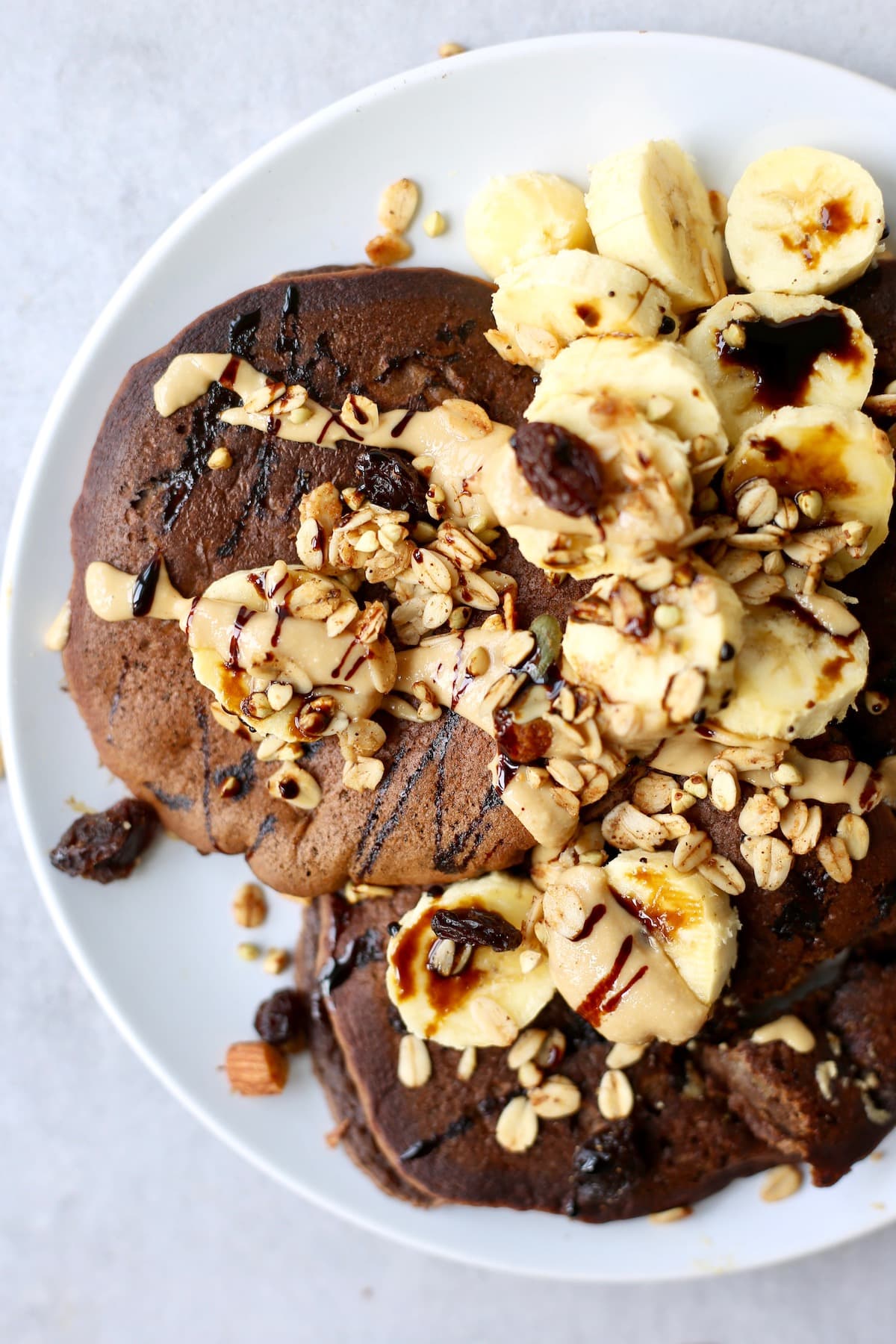 chocolate chickpea flour pancakes on a plate topped with sliced banana, nuts, raisins, granola chocolate syrup and nut butter