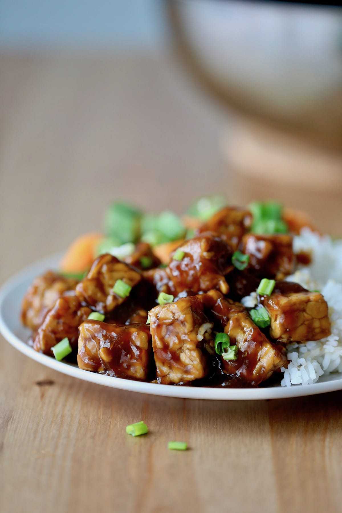 Sticky teriyaki tempeh with rice on a plate topped with vibrant sliced green onions. 