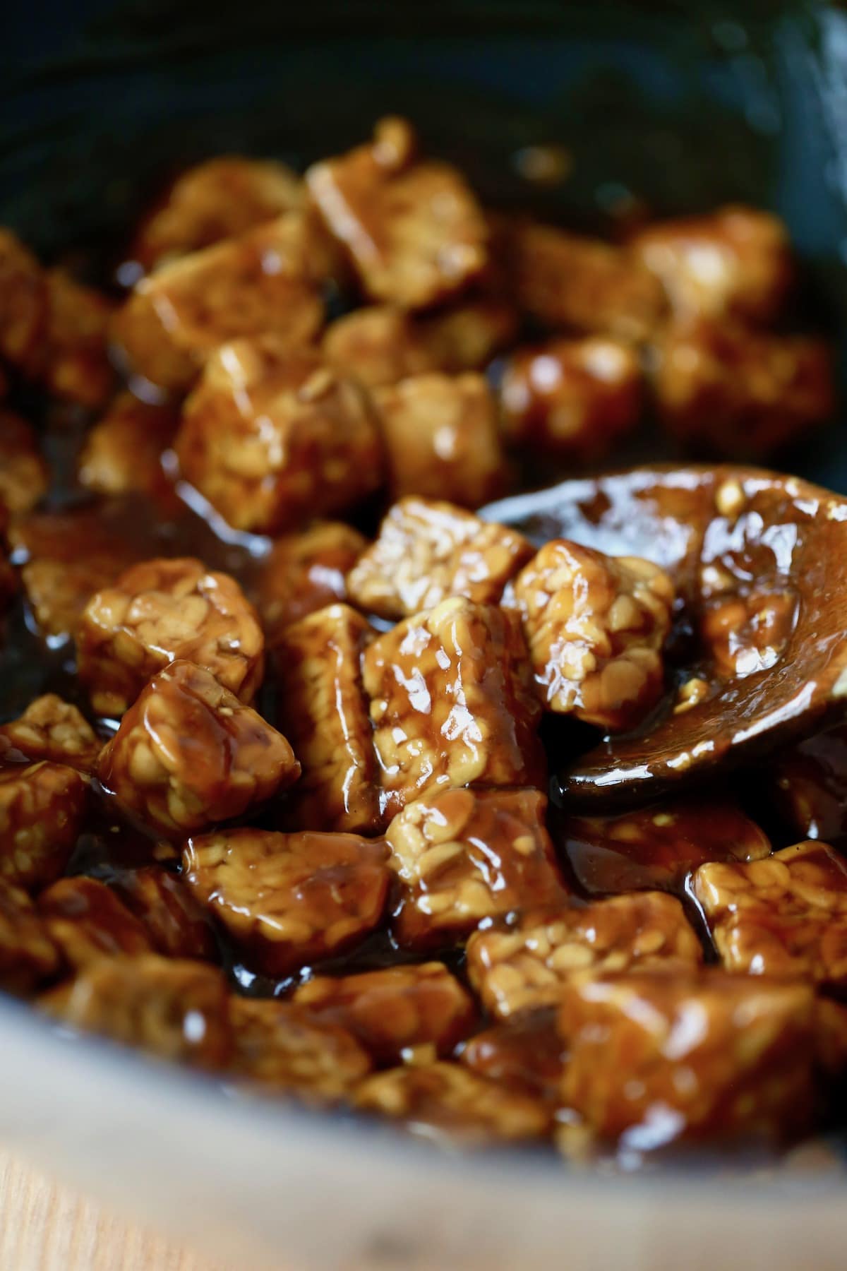 A wooden spoon stirring glossy teriyaki tempeh in a black wok. 