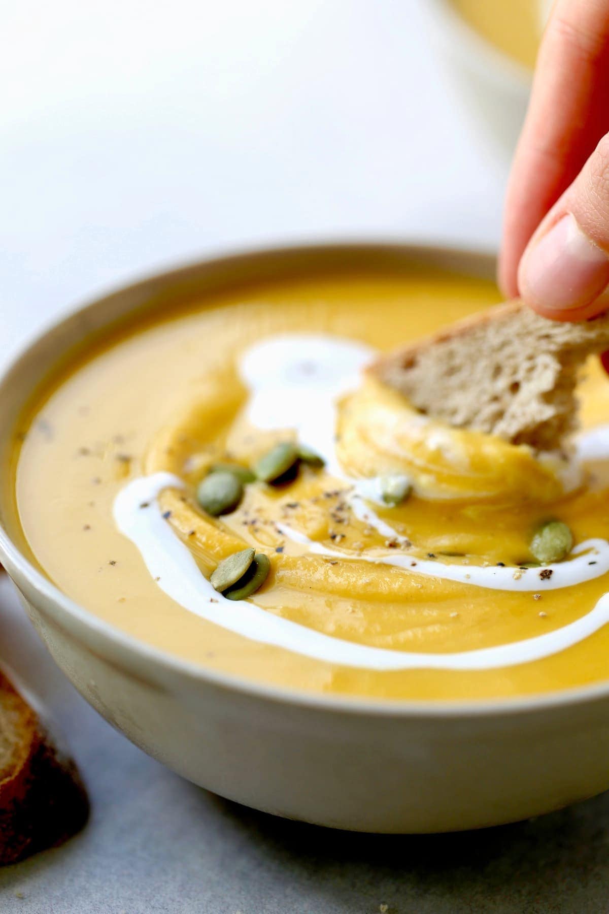 A close up of a hand using bread to scoop up some pumpkin peanut butter soup drizzled with coconut cream.