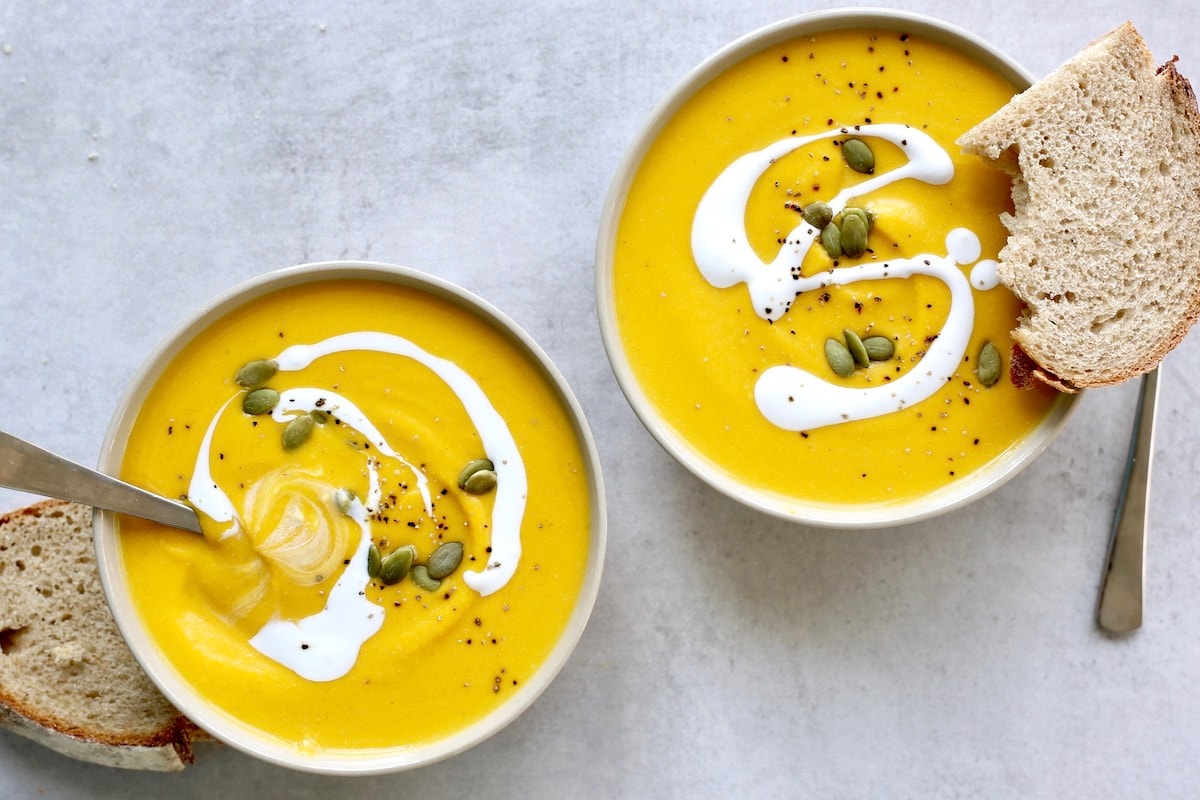 An overhead shot of two bowls of vegan pumpkin soup with coconut cream drizzled on top and bread on the side. 