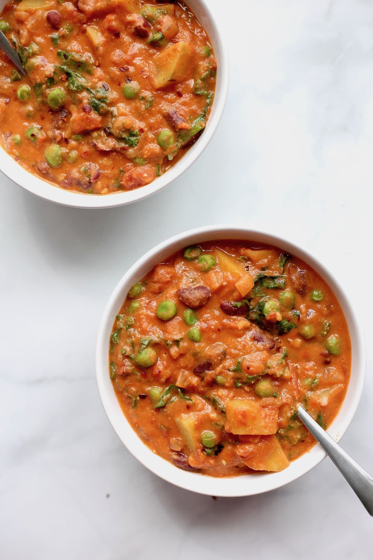 An overhead shot of two bowls of maafe full of peanut butter and tomato broth, green peas, sweet potatoes, kale and kidney beans.