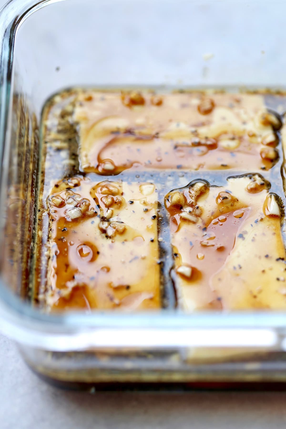 A 45 degree angle of tofu marinating in a glass dish with lots of fresh garlic, soy sauce and black pepper. 