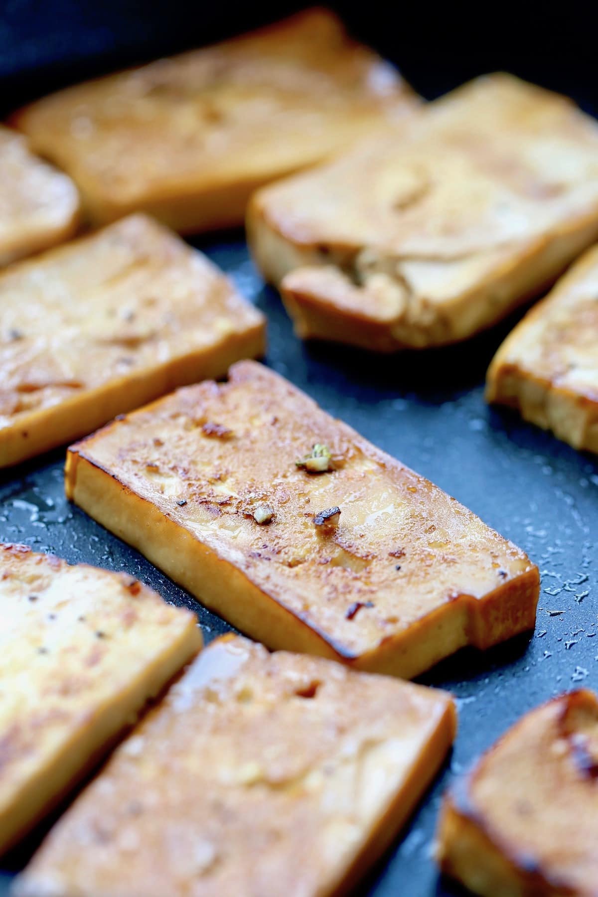 A rectangle of tofu browning on a black skillet with fresh garlic on top. 