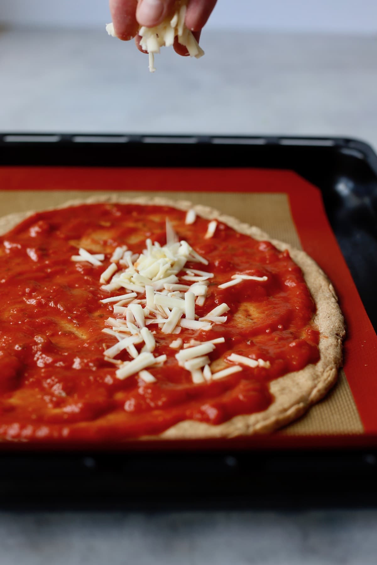 A hand sprinkling mozzarella cheese onto a pizza crust topped with marinara sauce. 