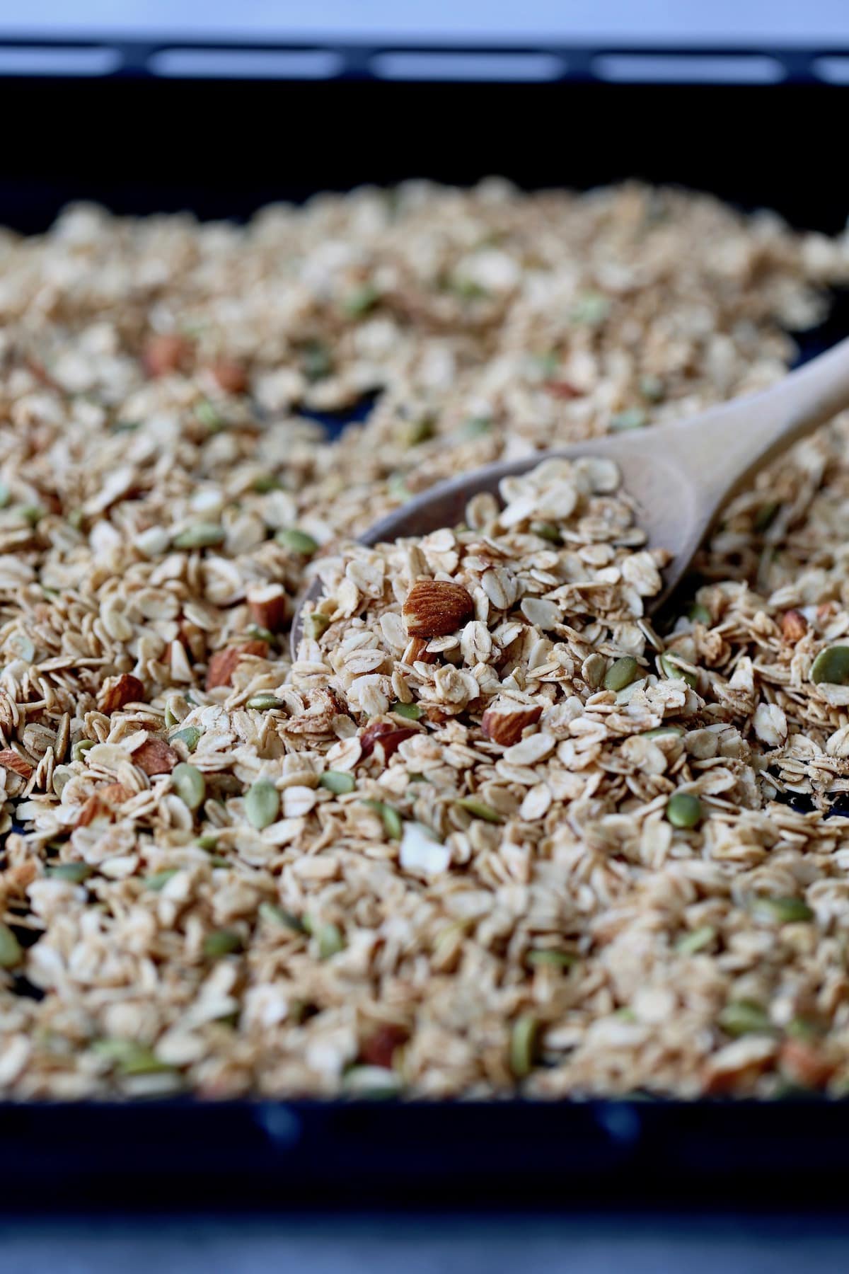 a large sheet pan of granola being scooped up with a wooden spoon