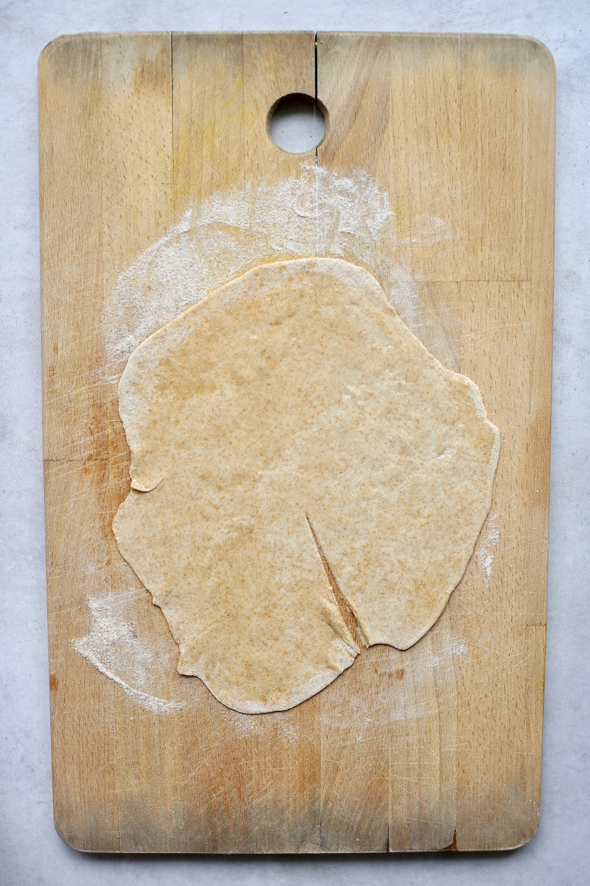 A thin circle of chapati dough rolled out on a floured wooden cutting board with a thin slice cut from the center to the edge. 