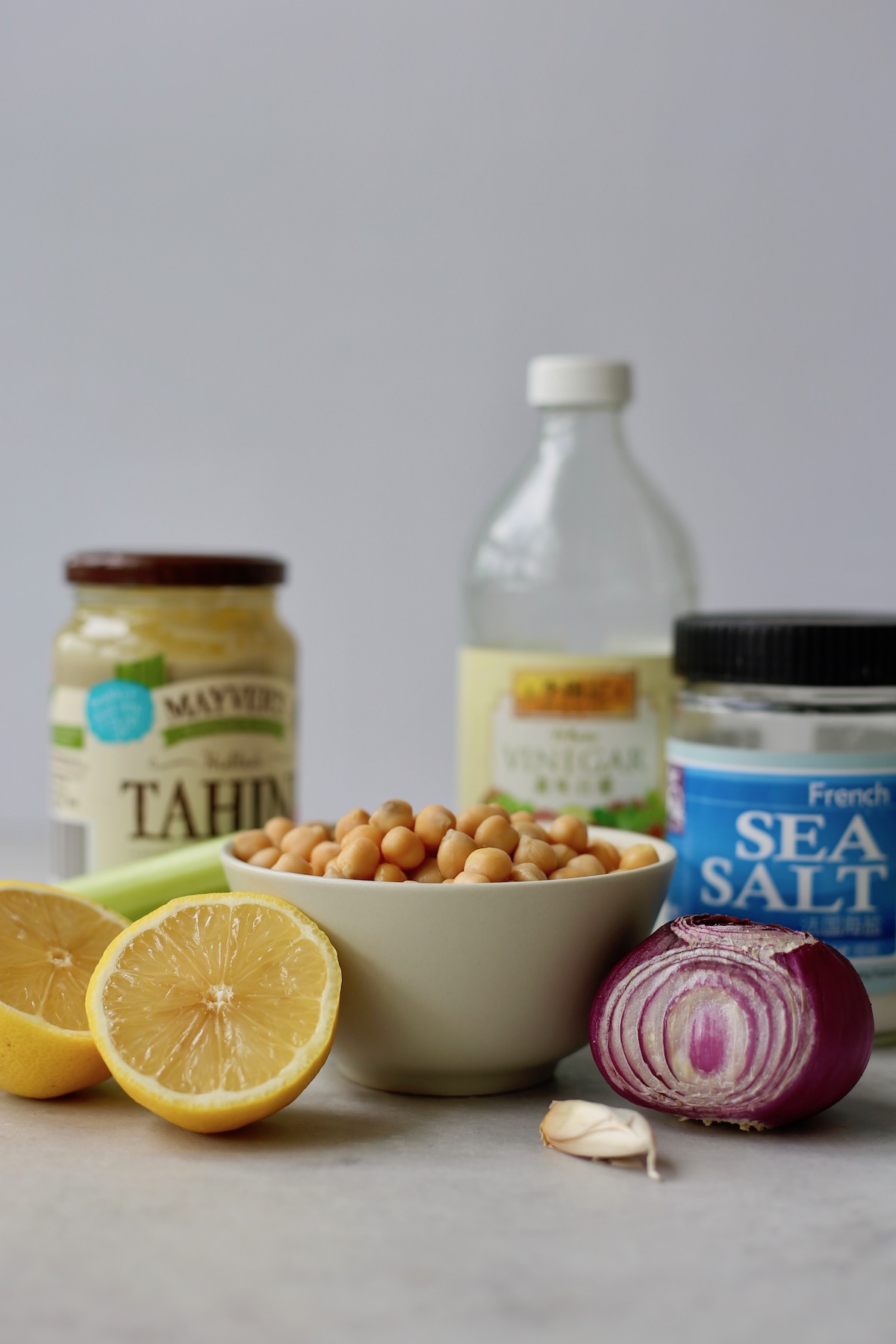 Tahini, vinegar, salt, red onion, lemons, chickpeas and celery on a table to make vegan chickpea salad. 