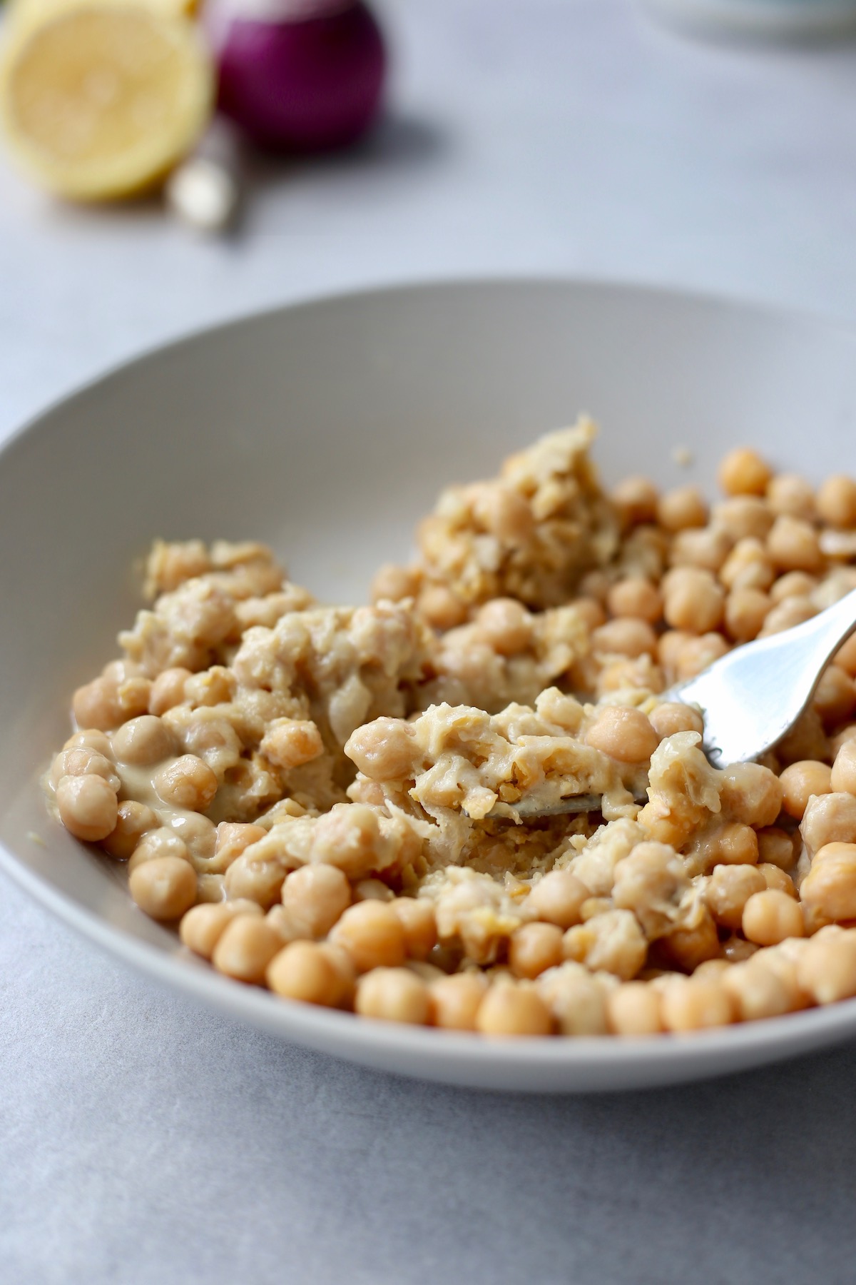 A fork mashing together chickpeas, tahini, lemon juice and vinegar in a wide bowl. 