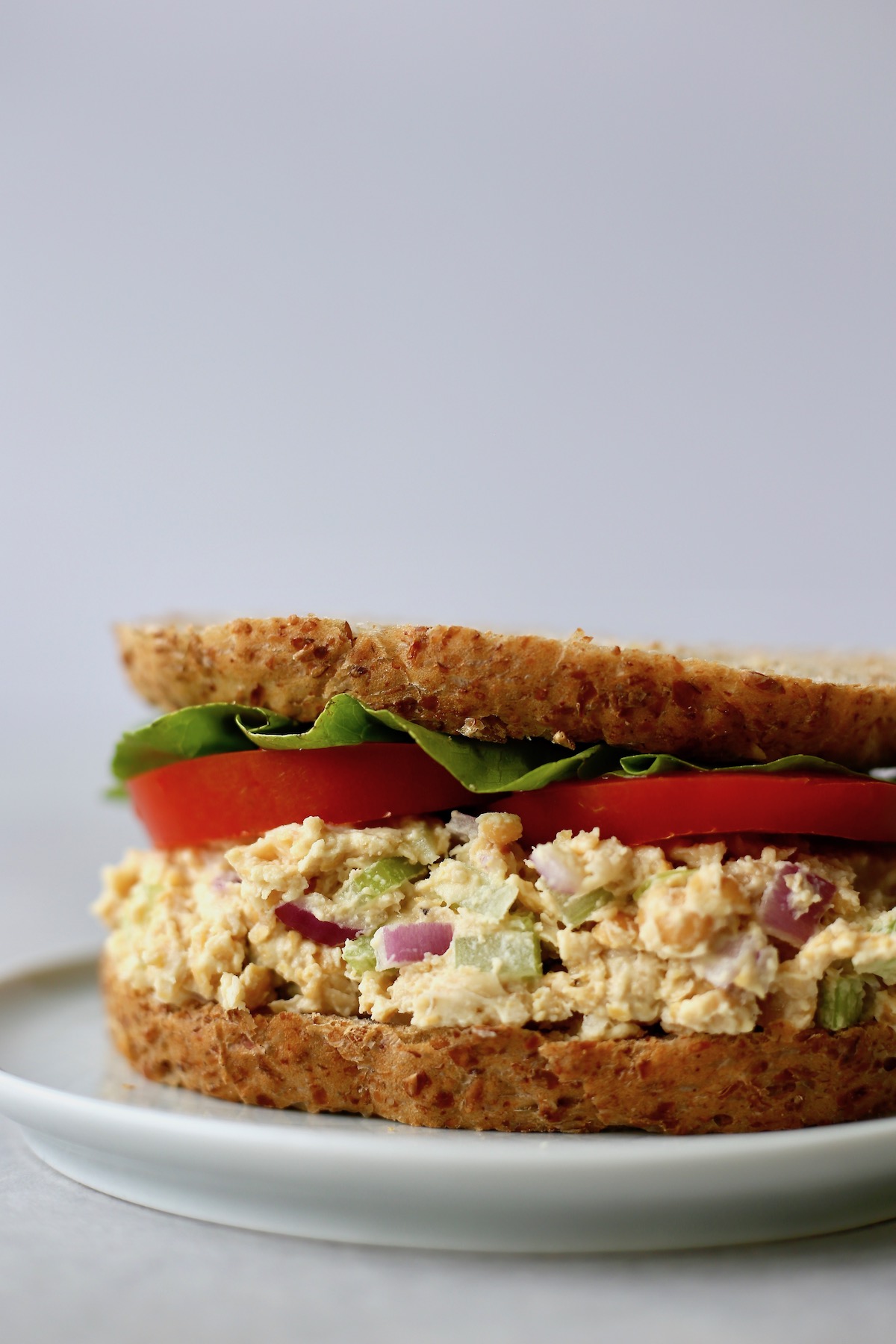 A sandwich filled with vegan chickpea salad, lettuce and tomato on a grey plate. 