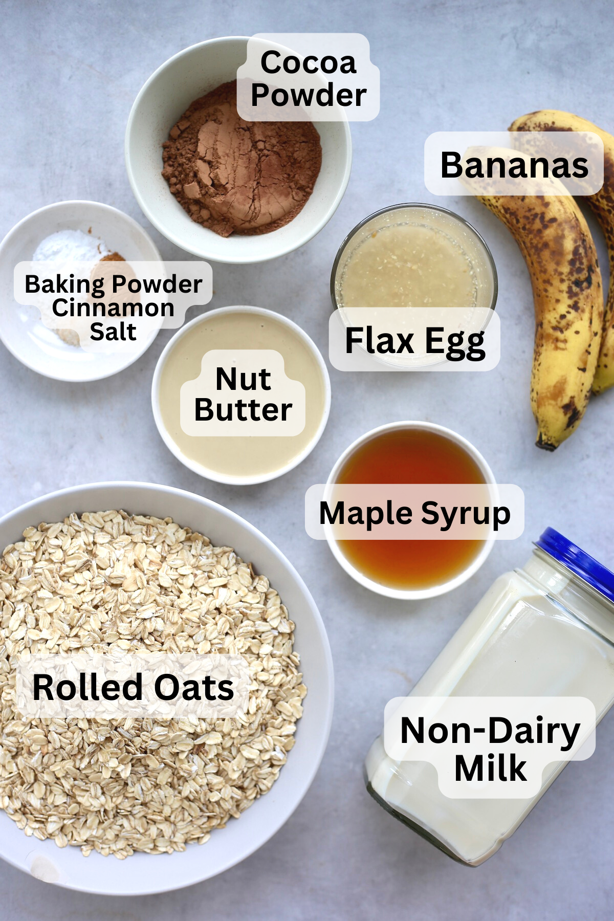 Cocoa powder, ripe bananas, rolled oats, maple syrup, non-dairy milk and other ingredients measured out into bowls on a table.