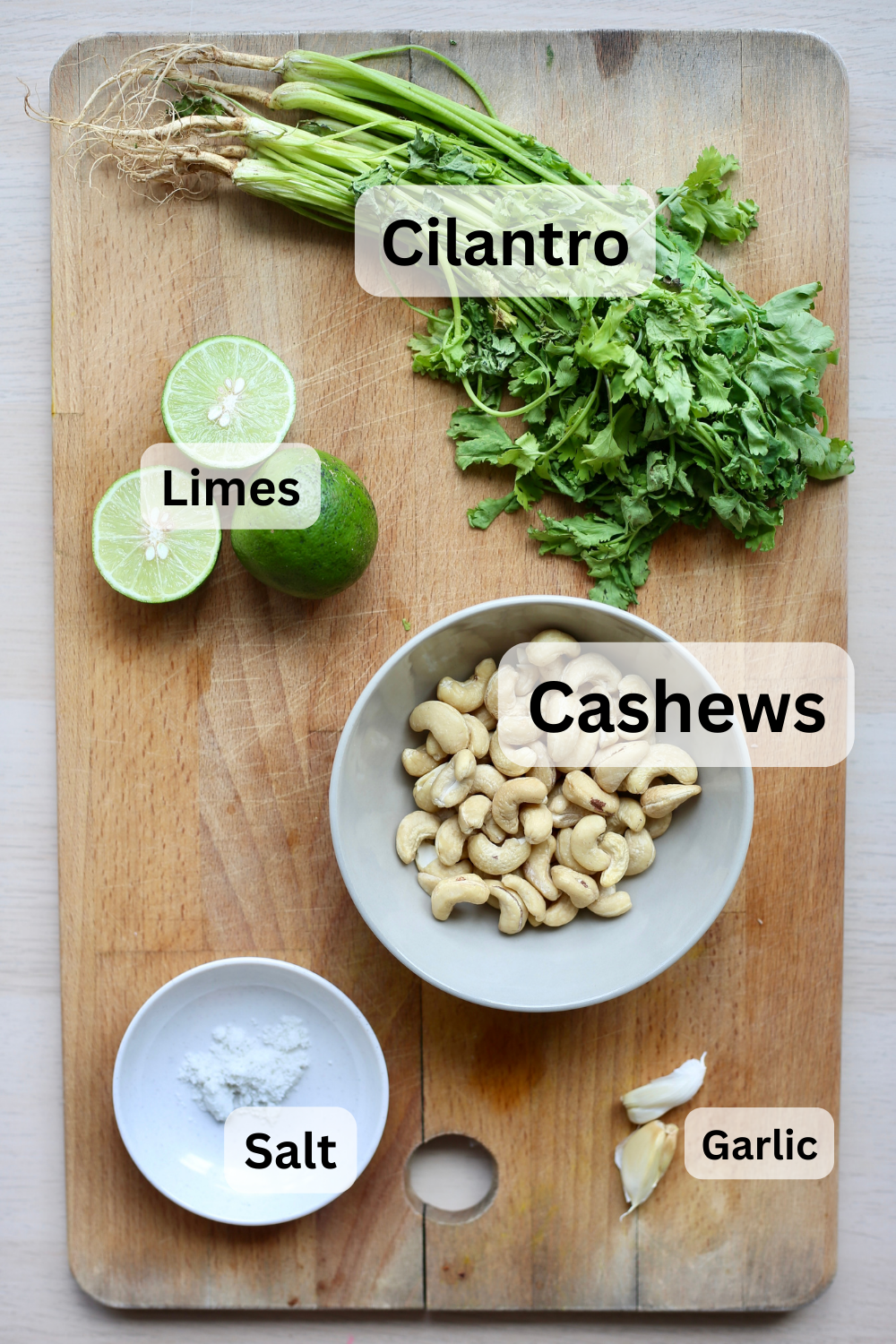 An overhead shot of cilantro, limes, cashews, garlic and salt laid out on a wooden cutting board. 