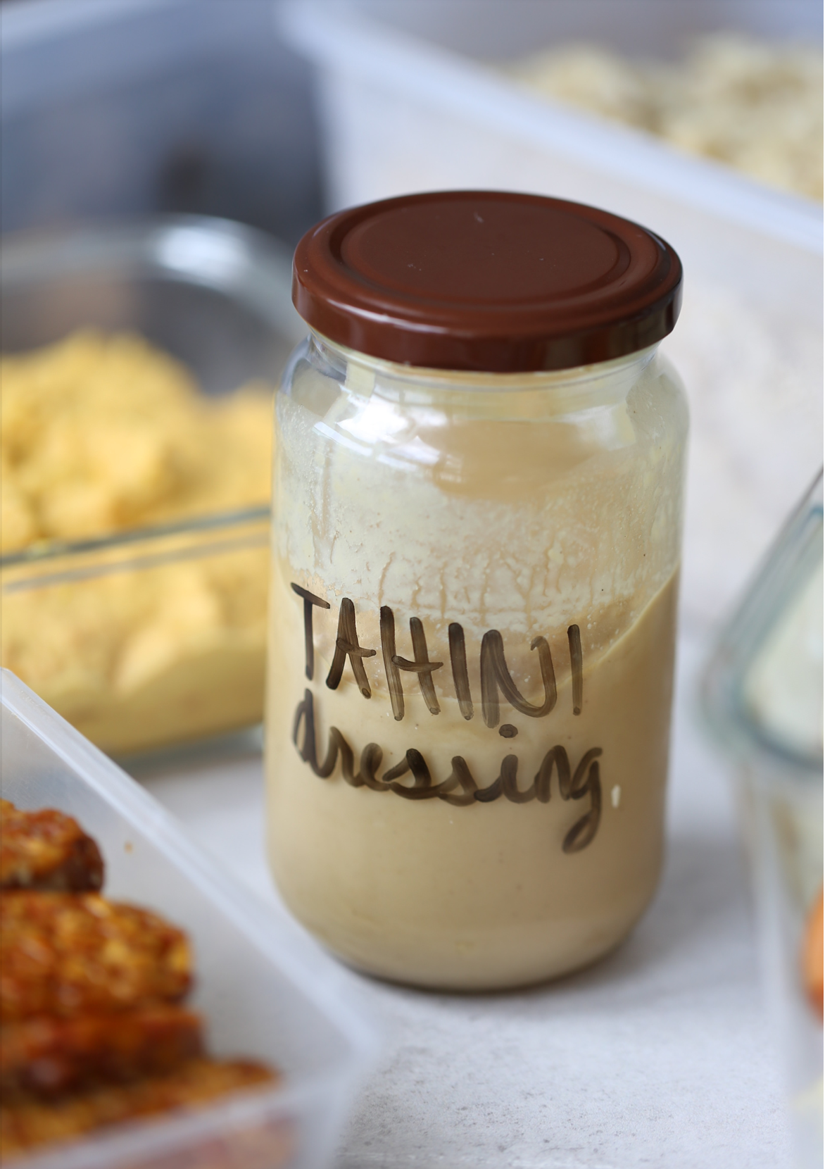 Prepared food with tahini soy dressing or crack sauce, in a glass jar with a brown lid and other dishes in the background. 