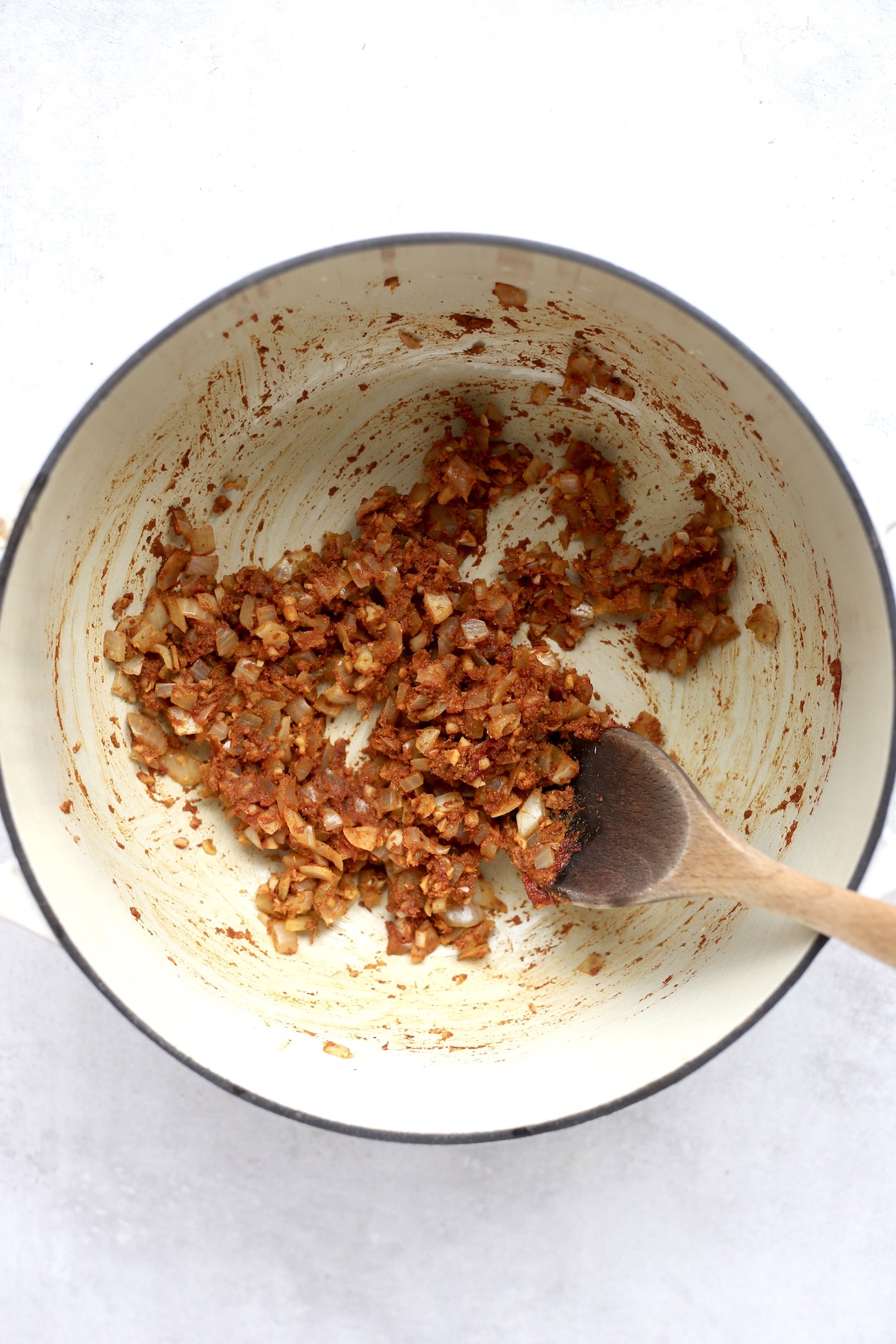 Red and brown spices coating onions and garlic in a large white pot with a wooden spoon inside. 