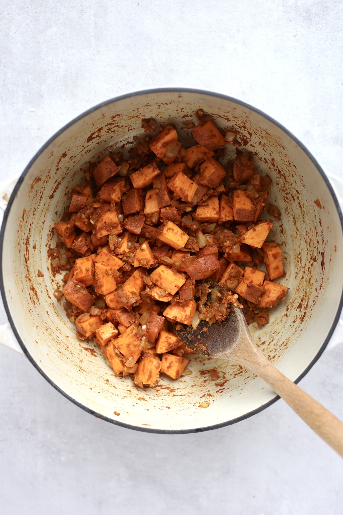 Chopped sweet potatoes coated in a red and brown spice mixture in a large white pot being stirred with a wooden spoon.