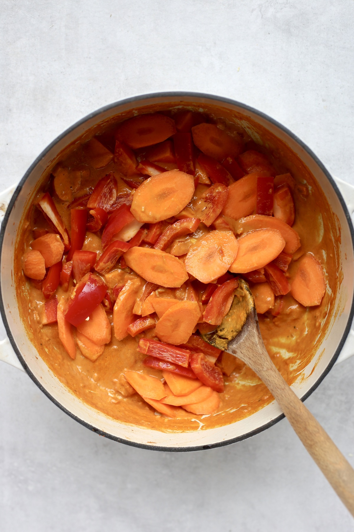 Sliced carrots and red bell pepper being stirred with a wooden spoon into an orange curry sauce in a white pot. 