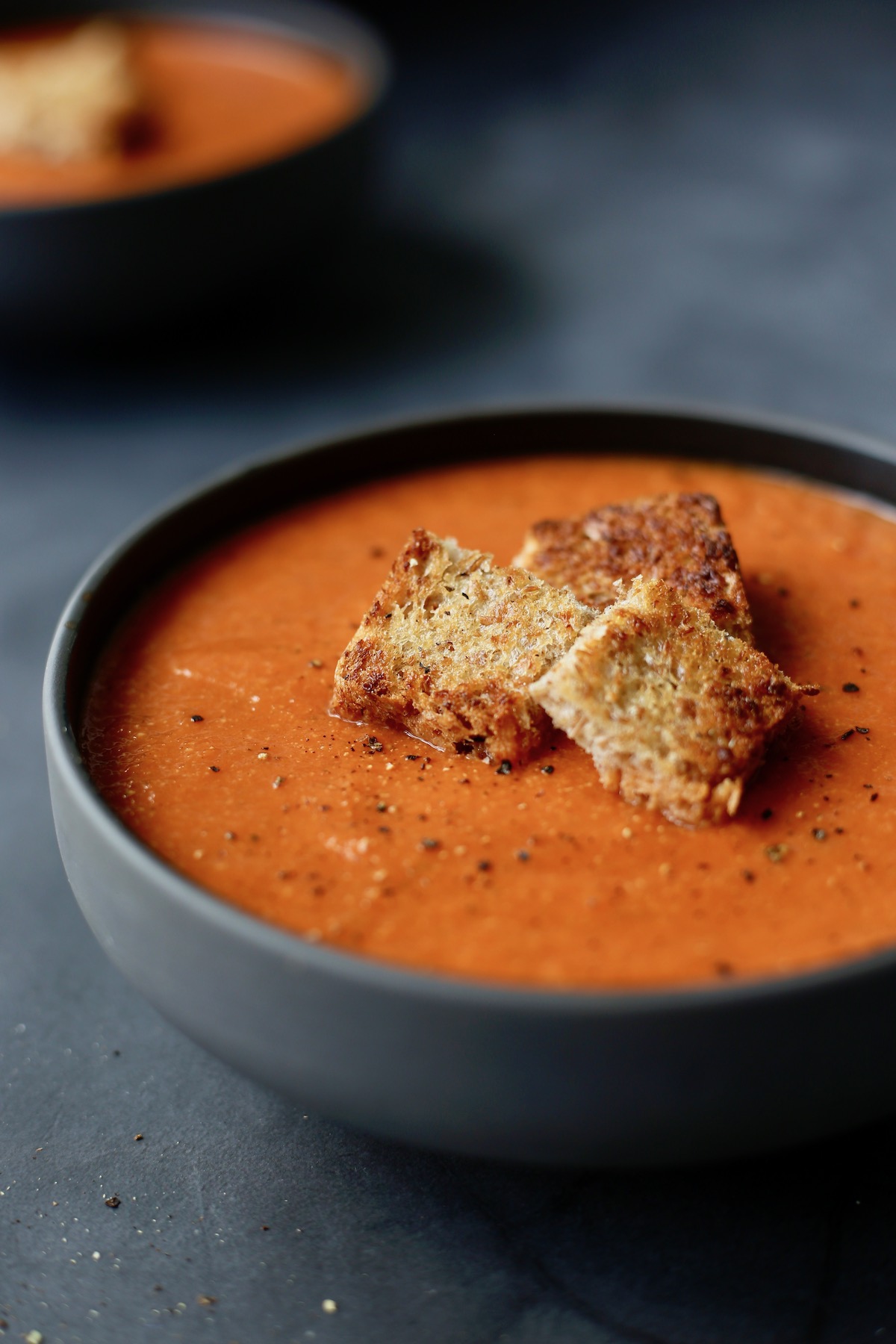 Creamy Tomato Zucchini Soup in a black bowl with golden brown croutons and black pepper on top.