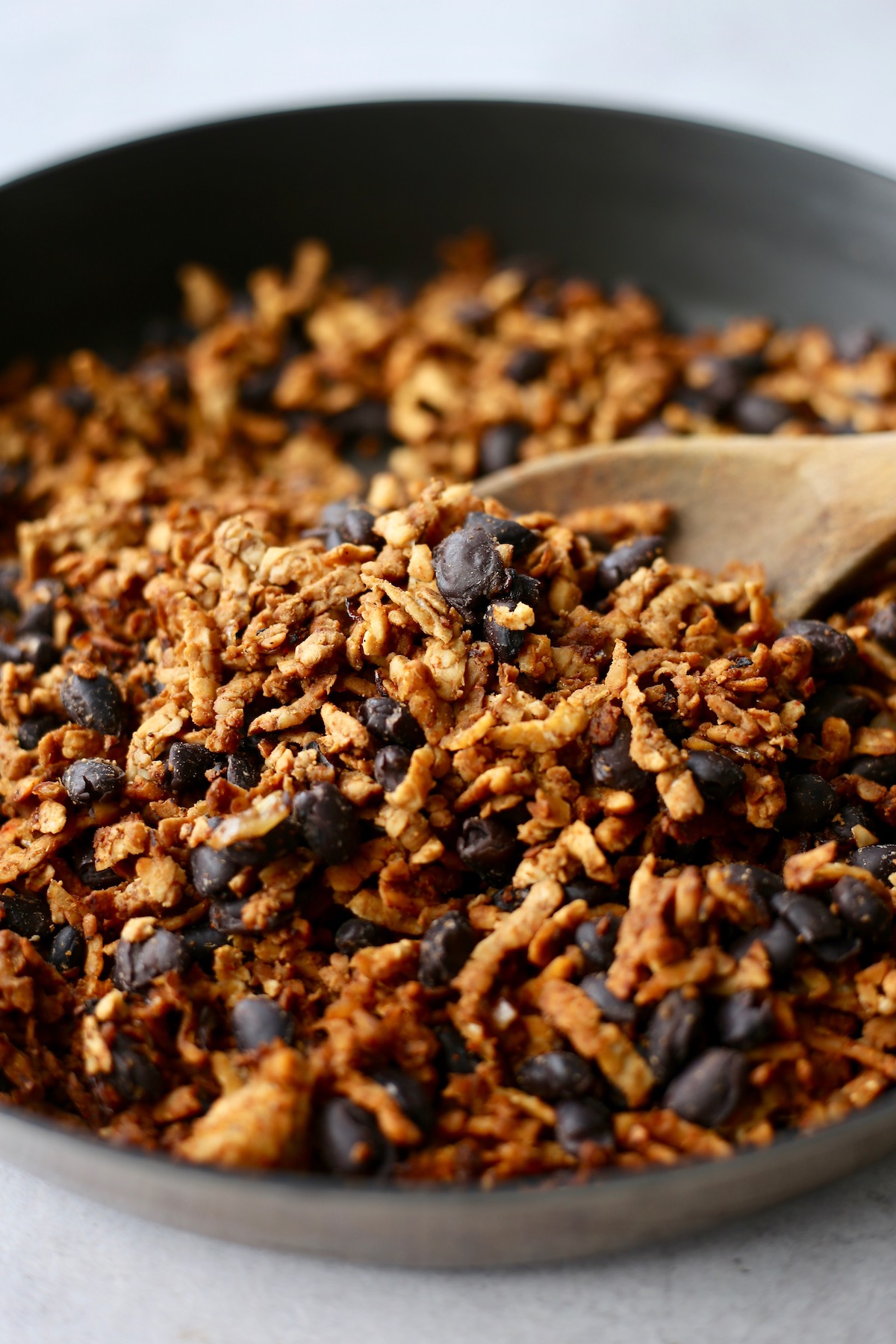 Tempeh and black beans coated in spices in a black skilled being stirred with a wooden spoon.