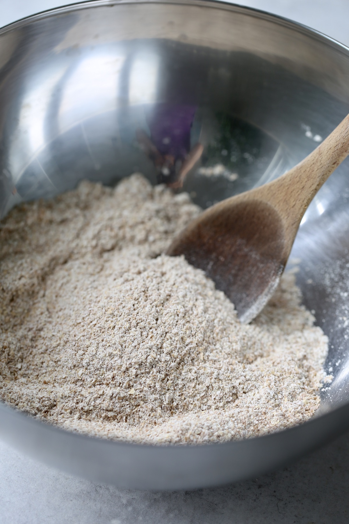 Flour and wheat bran being stirred together in a metal mixing bowl with a wooden spoon.