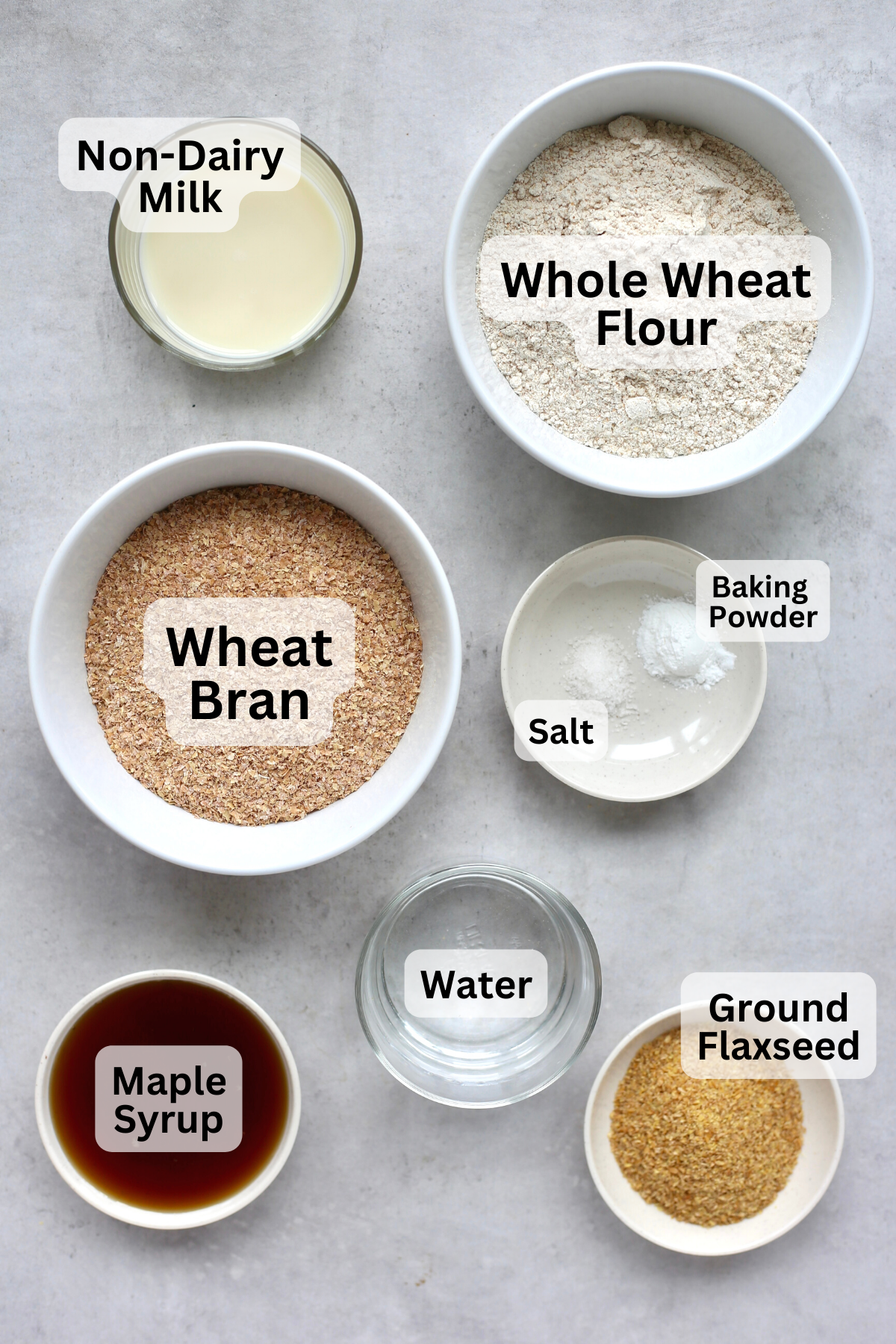 Wheat bran, whole wheat flour, ground flaxseed, maple syrup and other ingredients measured out into bowls with labels overtop.