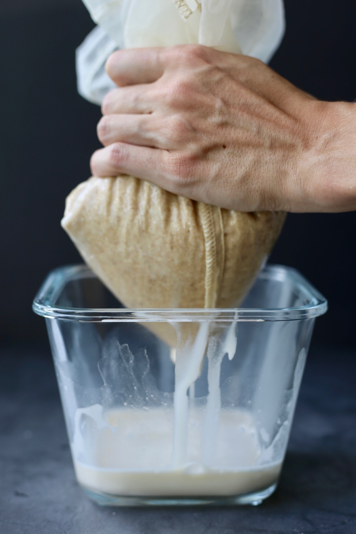A hand squeezing oat milk out of a nut milk bag into a square glass container.