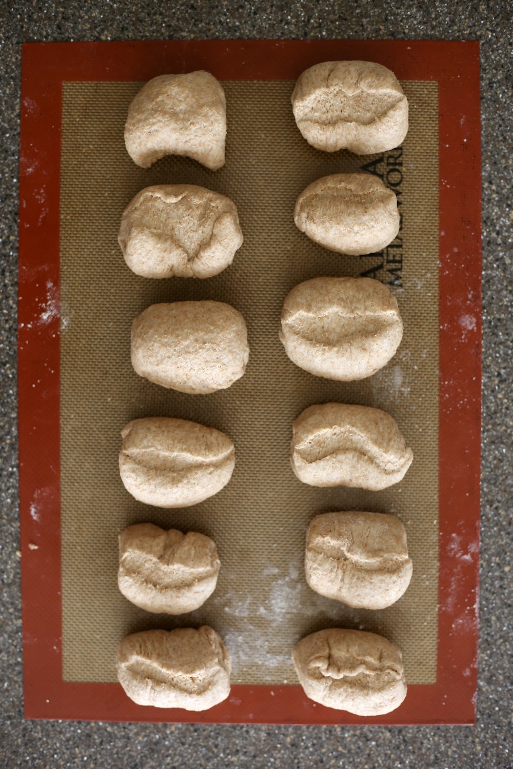 Twelve pieces of whole wheat bagel dough resting on a silicone baking mat.