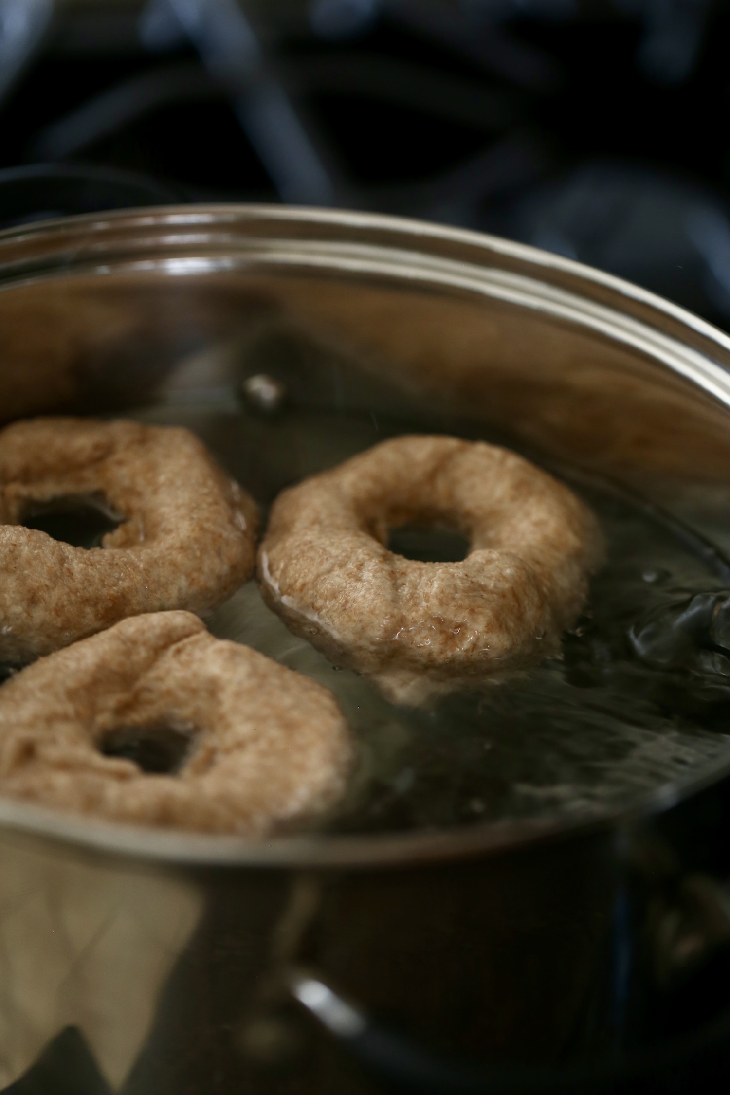 Three whole wheat bagels boiling in a large silver pot of water.