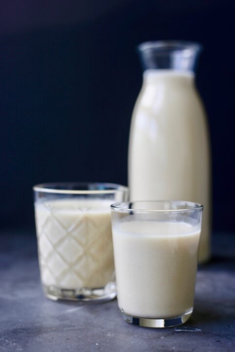 two glasses of oat milk next to a jar of oat milk