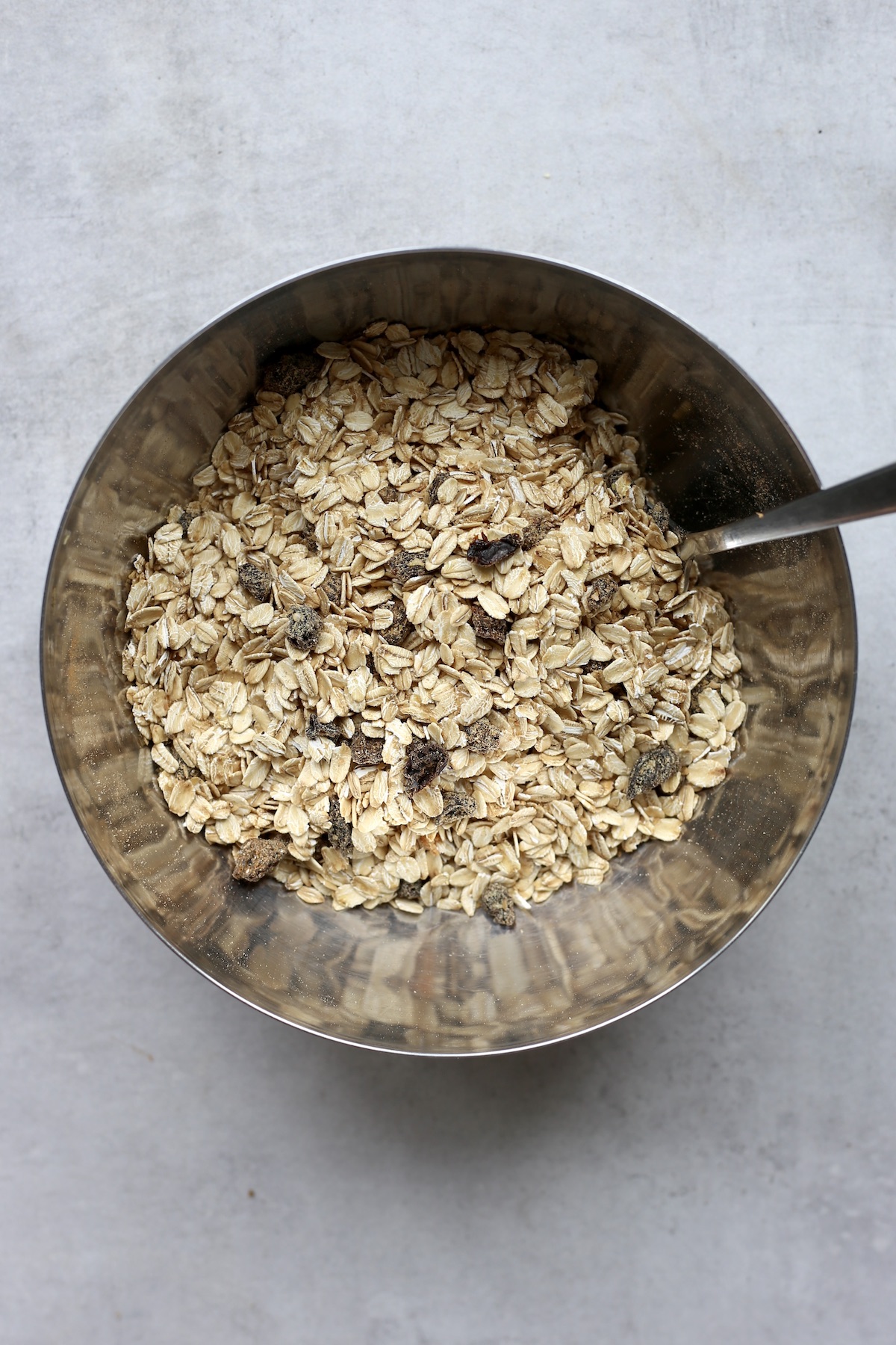 Rolled oats, raisins, ground flaxseed and spices being stirred together in a metal mixing bowl.