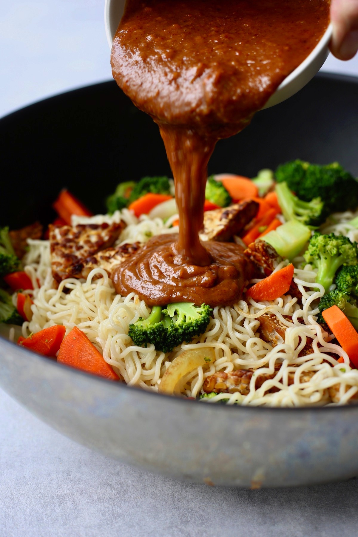 peanut satay sauce being poured over a bowl of noodles, tempeh and veggies