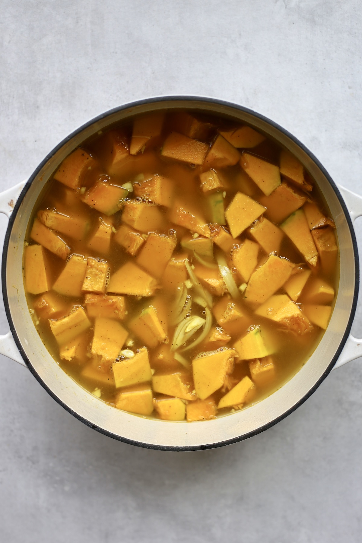 Large pieces of pumpkin and sliced onion in lots of broth in a white dutch oven. 