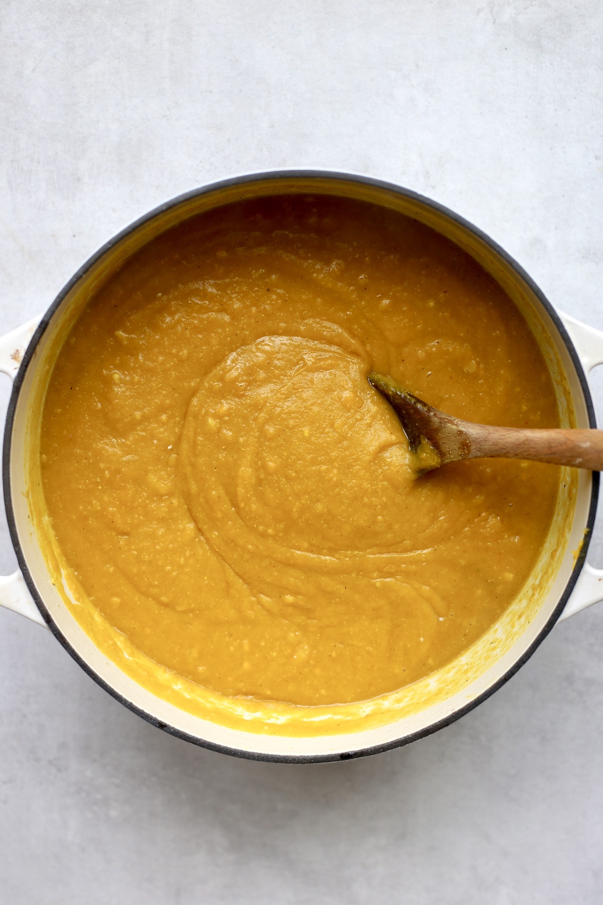 A wooden spoon stirring creamy pumpkin peanut butter soup in a large white dutch oven. 
