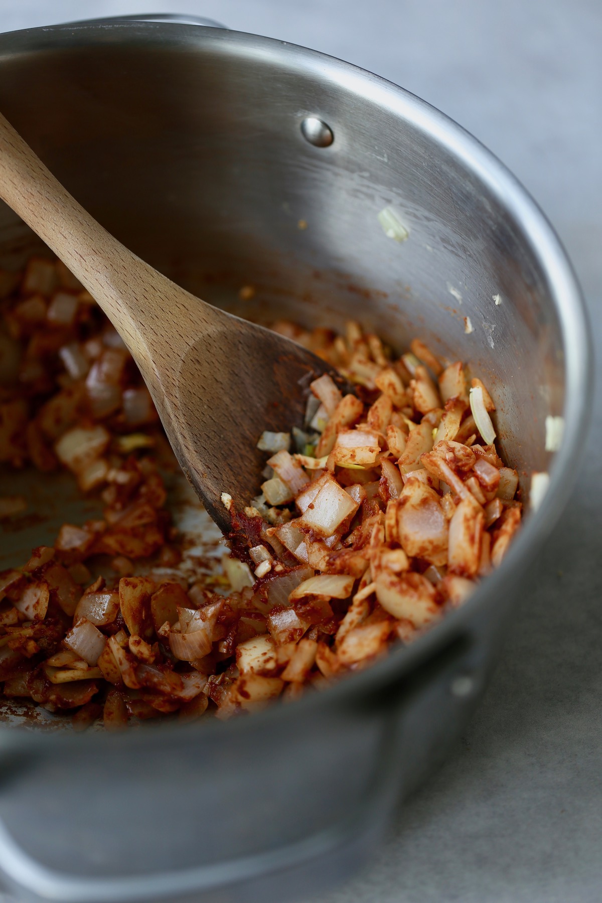 Onions and garlic coated in tomato paste and spices being stirred with a wooden spoon in a silver pot.