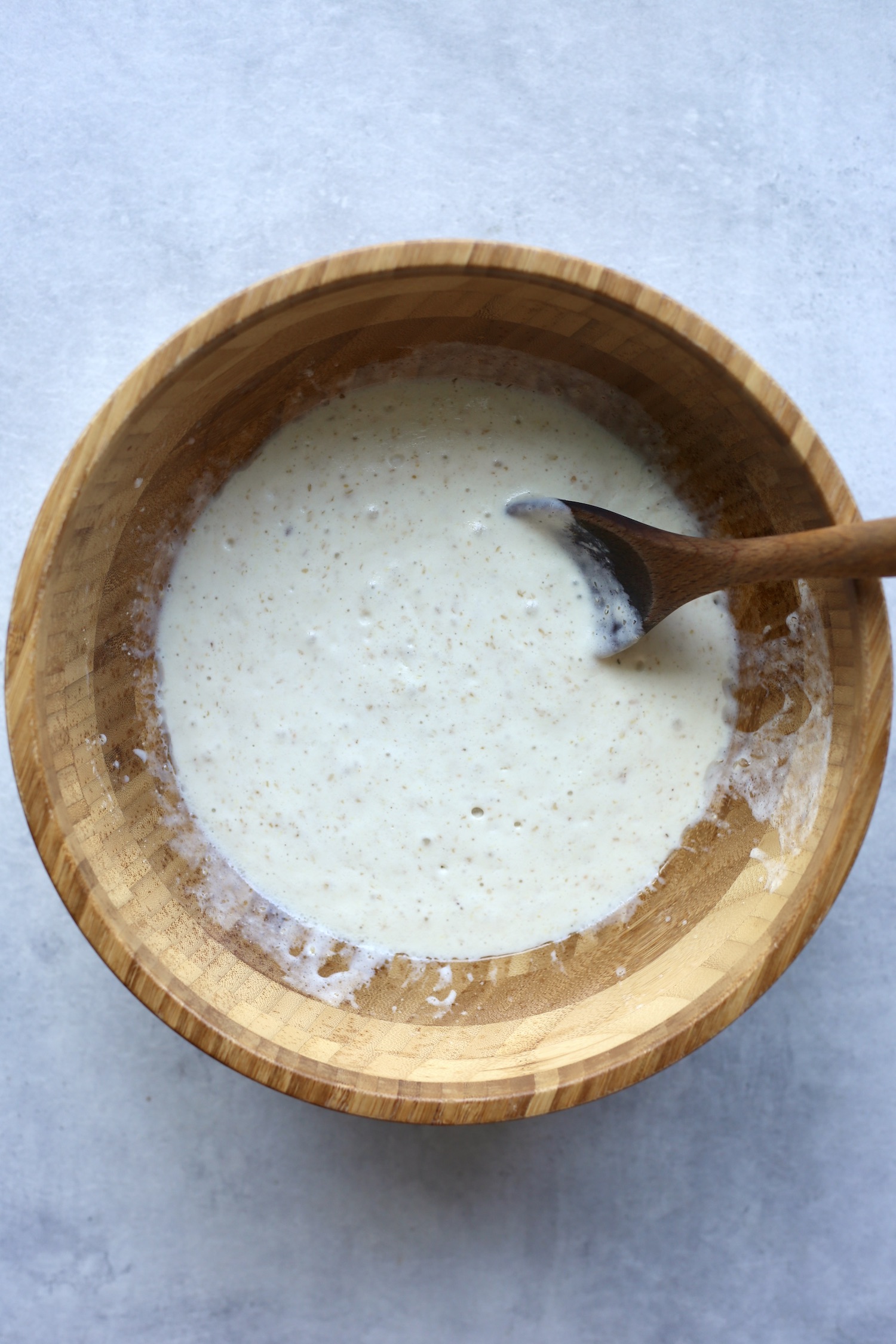 A wooden spoon stirring sourdough muffin batter in a large wooden mixing bowl. 