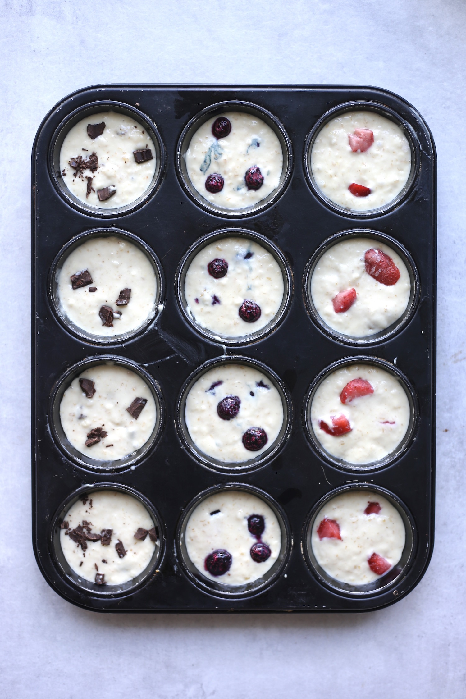 Vegan sourdough muffin batter in a muffin tin before baking topped with chocolate chips, blueberries and strawberries. 