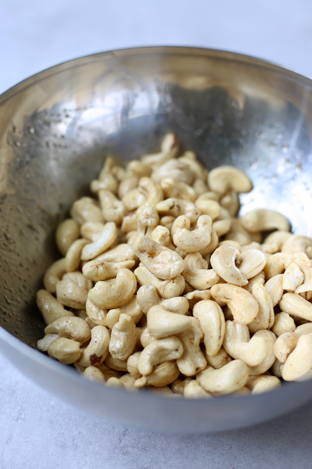 Raw cashews being coated with olive oil, maple syrup and spices in a mixing bowl.